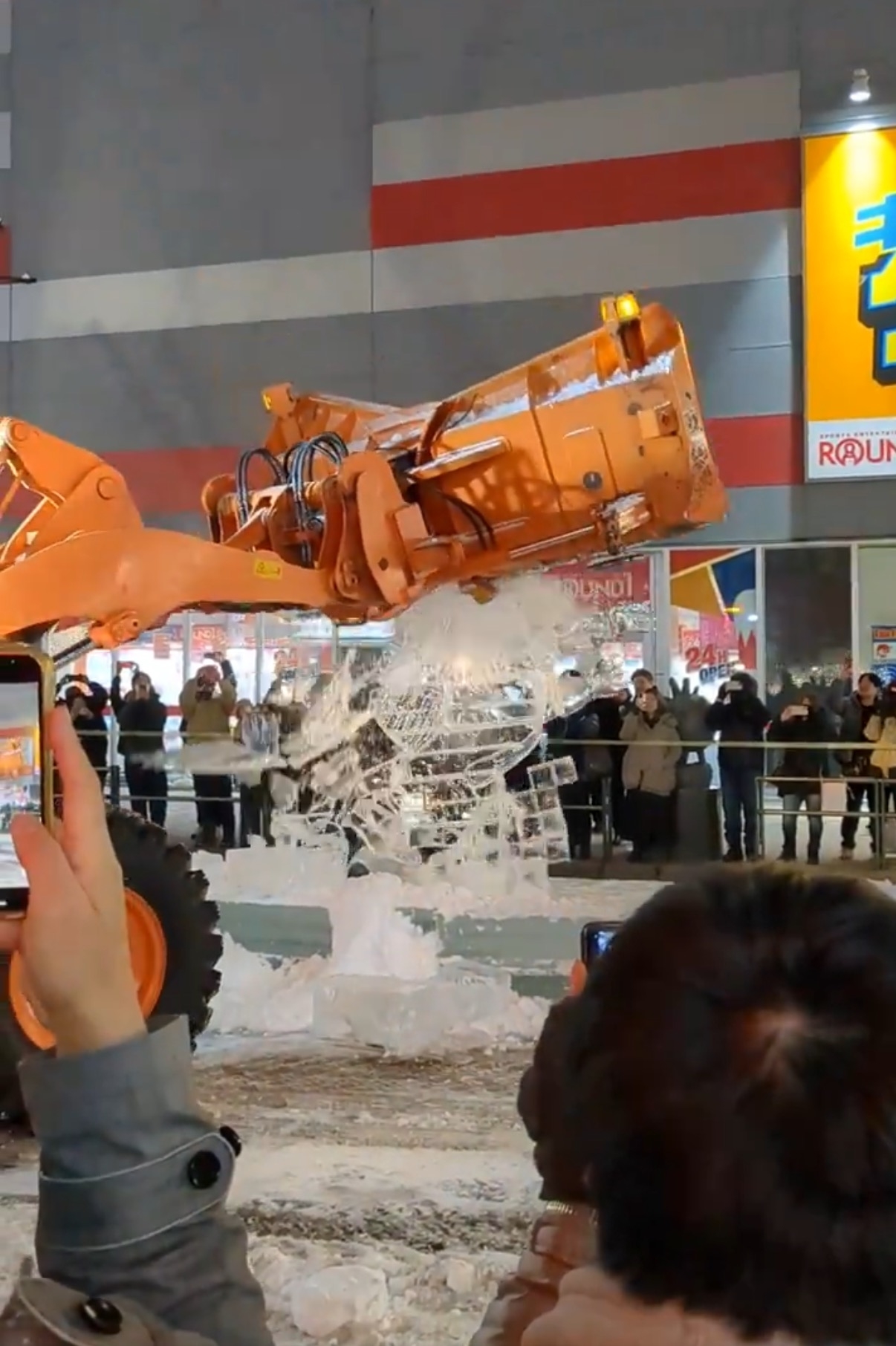 オレンジ色の建設用ショベルが氷の彫刻を破壊している様子。見物人が周囲にいる。