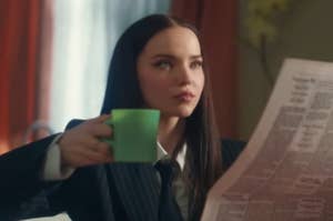 Dove Cameron in a suit, holding a coffee cup and reading a newspaper.