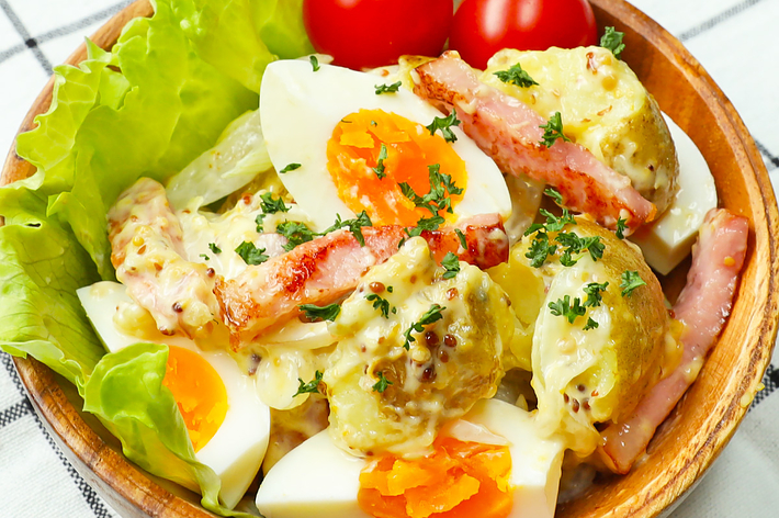 木のボウルに入ったポテトサラダ、ゆで卵、ベーコン、レタスとトマトが添えられている。