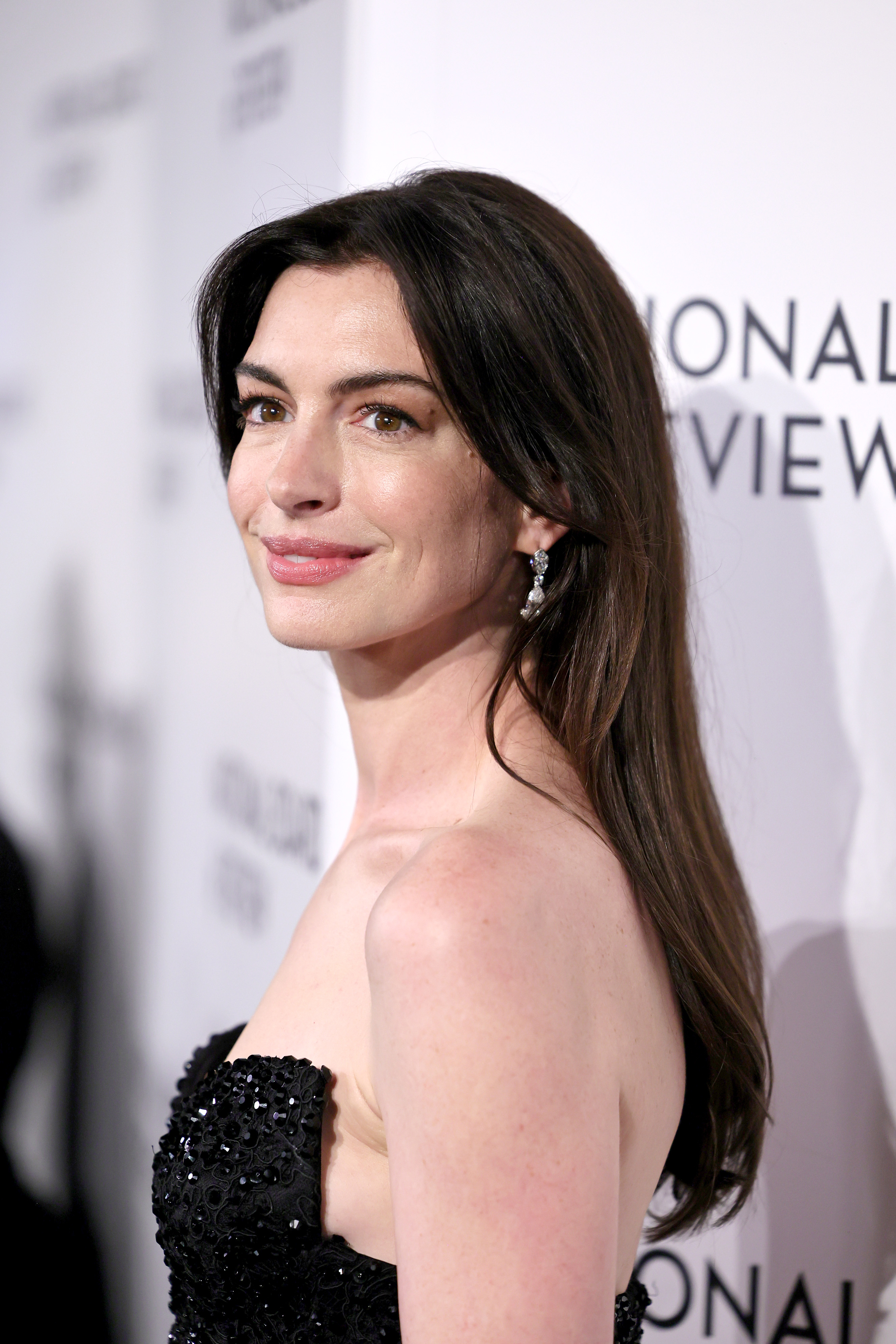 Anne Hathaway in a black sequined dress with one shoulder design, posing with a smile at an event
