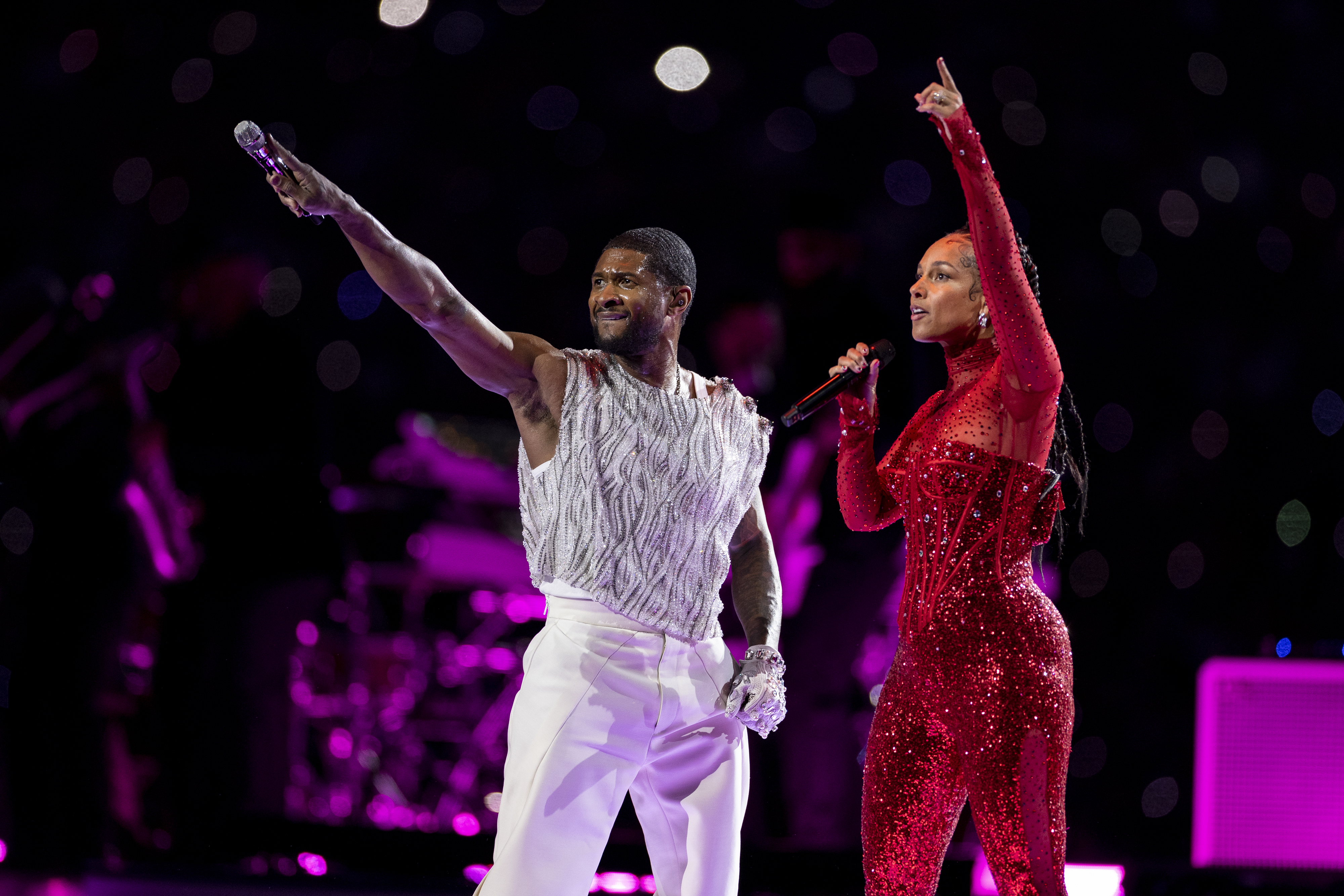 Usher and Alicia performers on stage, one in a fringed white top and white pants, the other in a sparkling red bodysuit, both with microphones