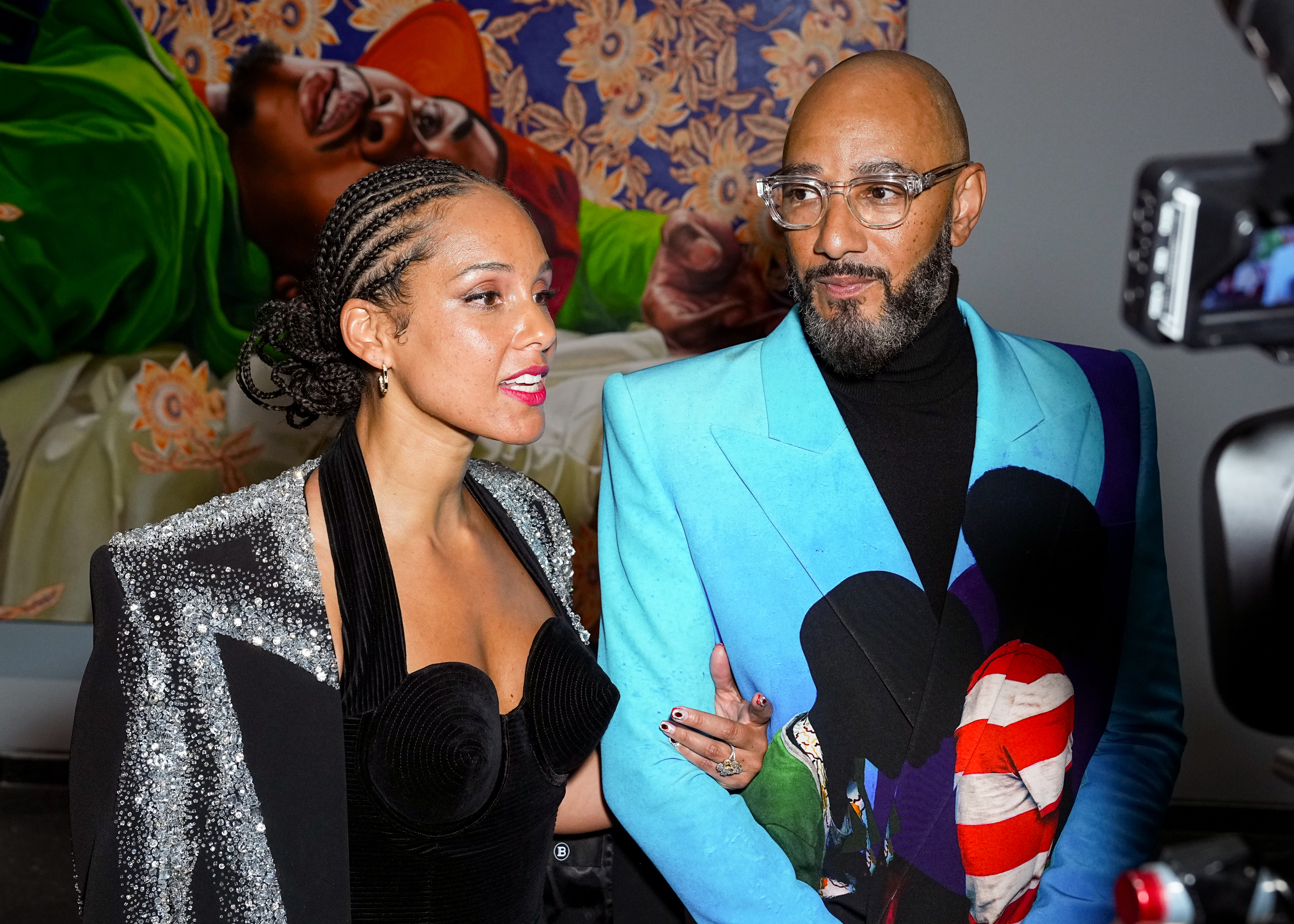 Alicia and Swizz Beatz people stand together; one in a sequined outfit, the other in a blue blazer with a graphic t-shirt, before artwork