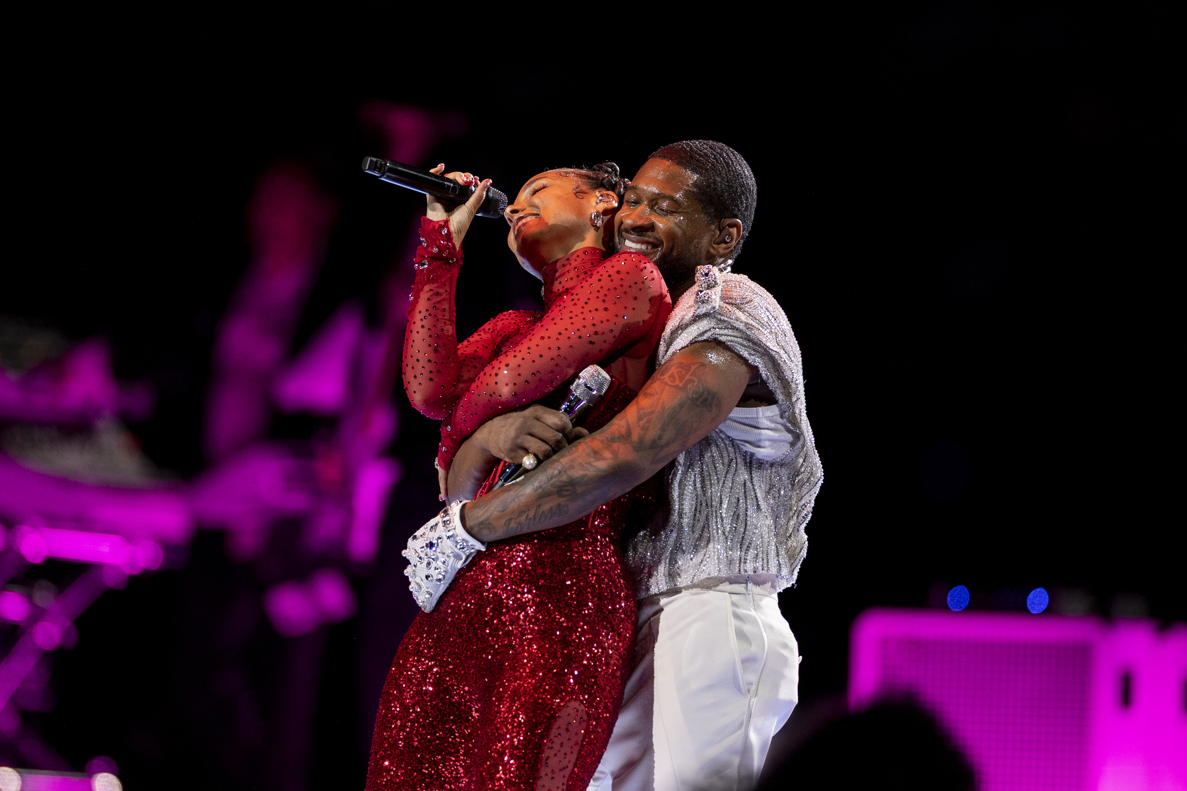 Usher hugging Alicia performers embracing while one holds a microphone, both in sparkly stage outfits