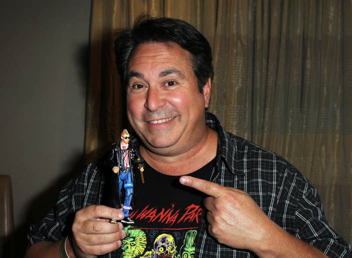 Brian Peck holding a small figurine, wearing plaid shirt and graphic tee, smiling at the camera