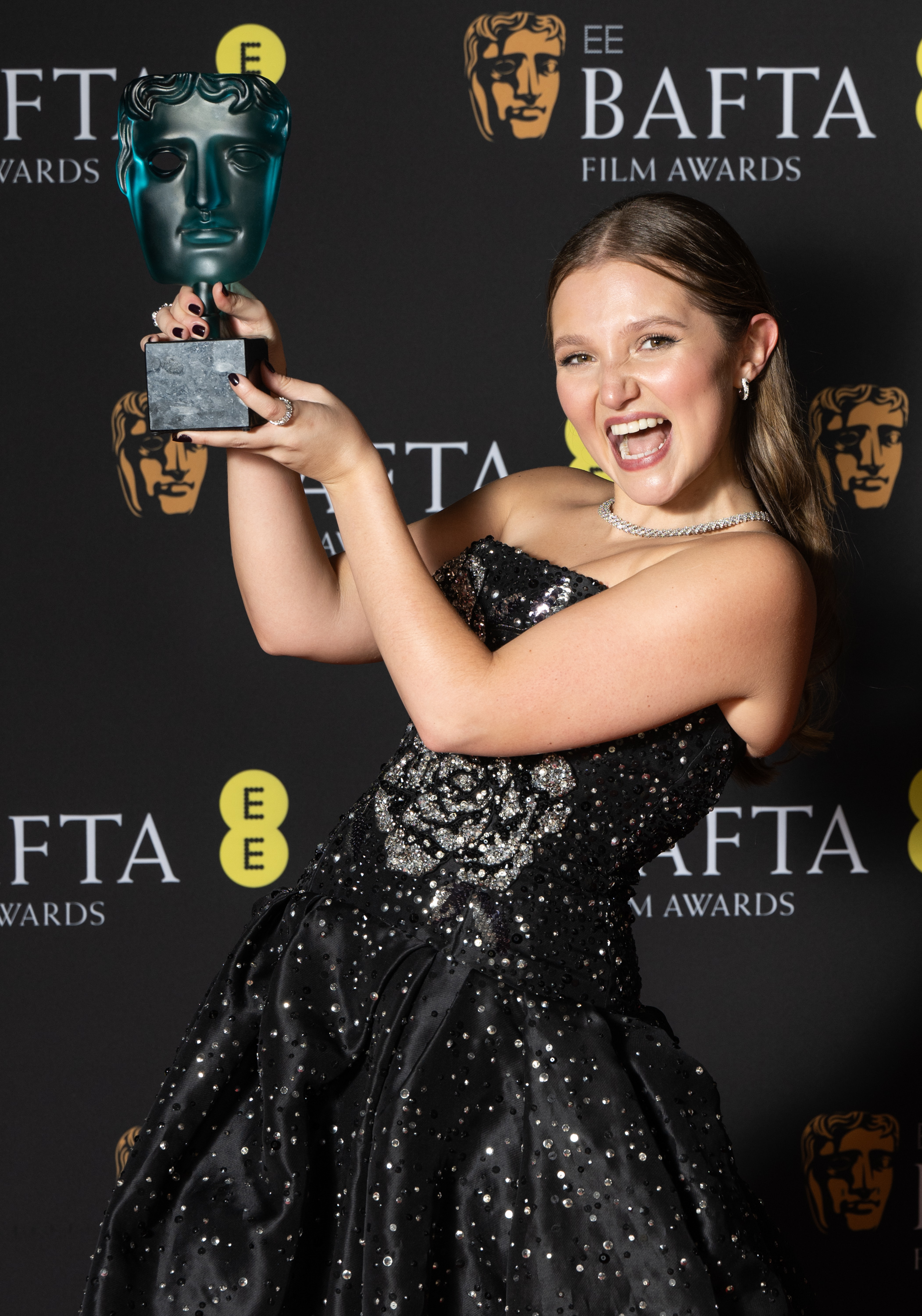 Woman in sparkling strapless gown taking a selfie with a BAFTA award