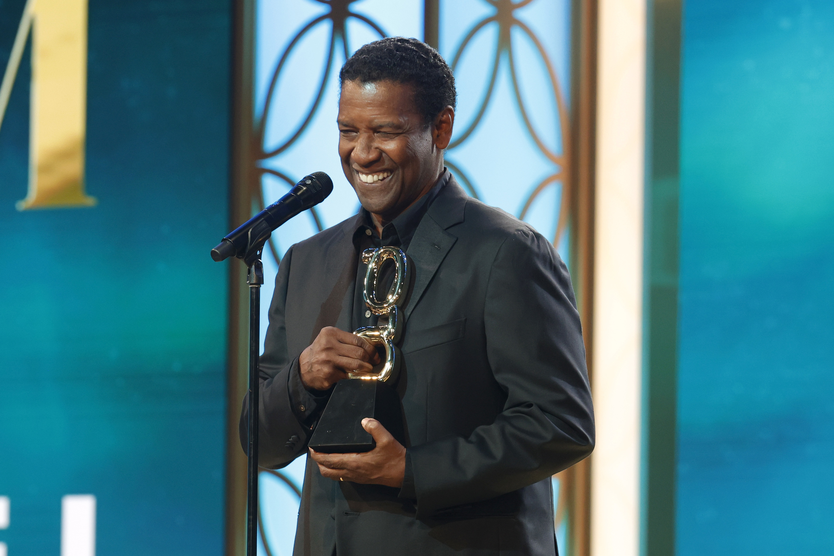Denzel Washington smiling, holding a trophy at a podium with a microphone