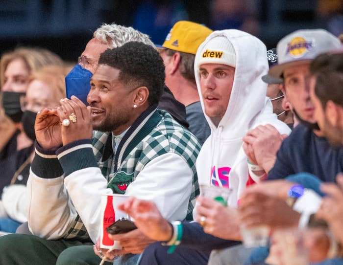 Usher and Justin Bieber sitting courtside at a basketball game. Usher wears a checkered jacket; Bieber is in a hoodie