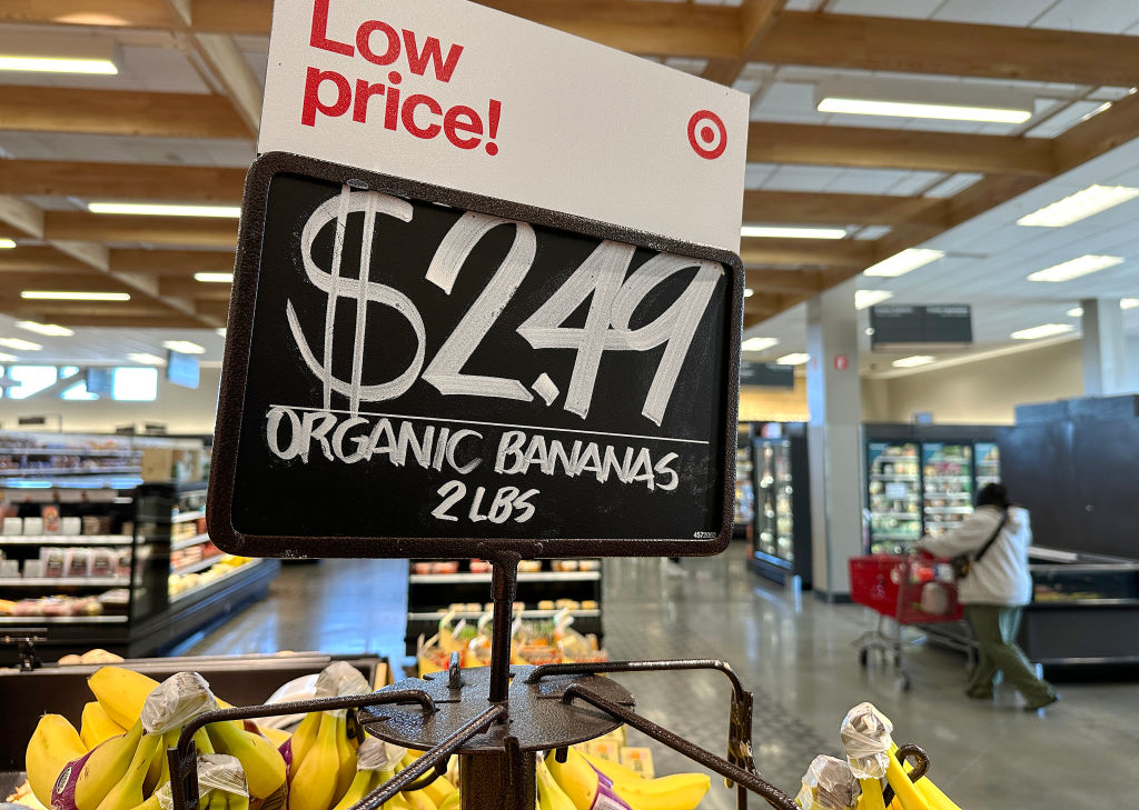 A Target price sign reads &quot;$2.49 ORGANIC BANANAS 2 lbs&quot; hanging above bananas in a store; there&#x27;s a shopper with a cart in the background