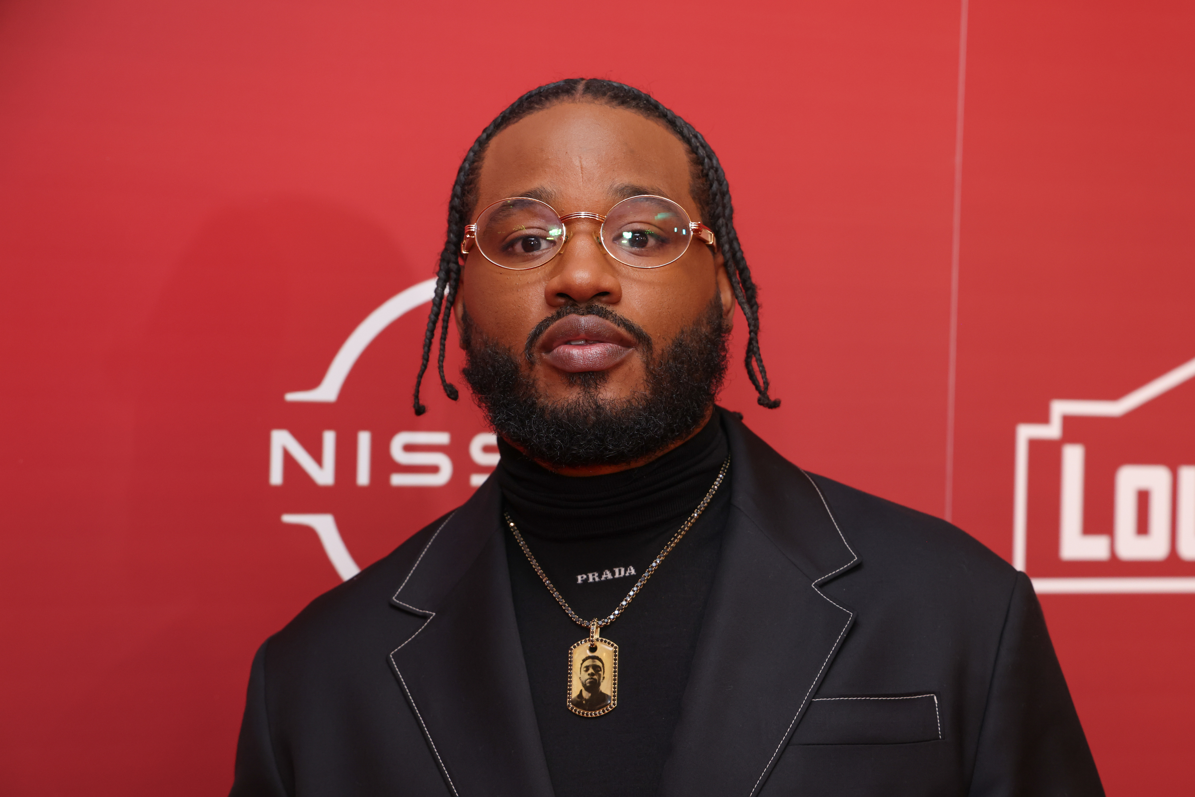 Man with braided hair, black turtleneck and jacket, Prada necklace, posing against a red background