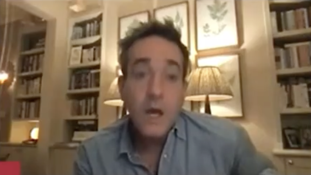 Matthew in a blue shirt speaking during a video call with bookshelves in the background