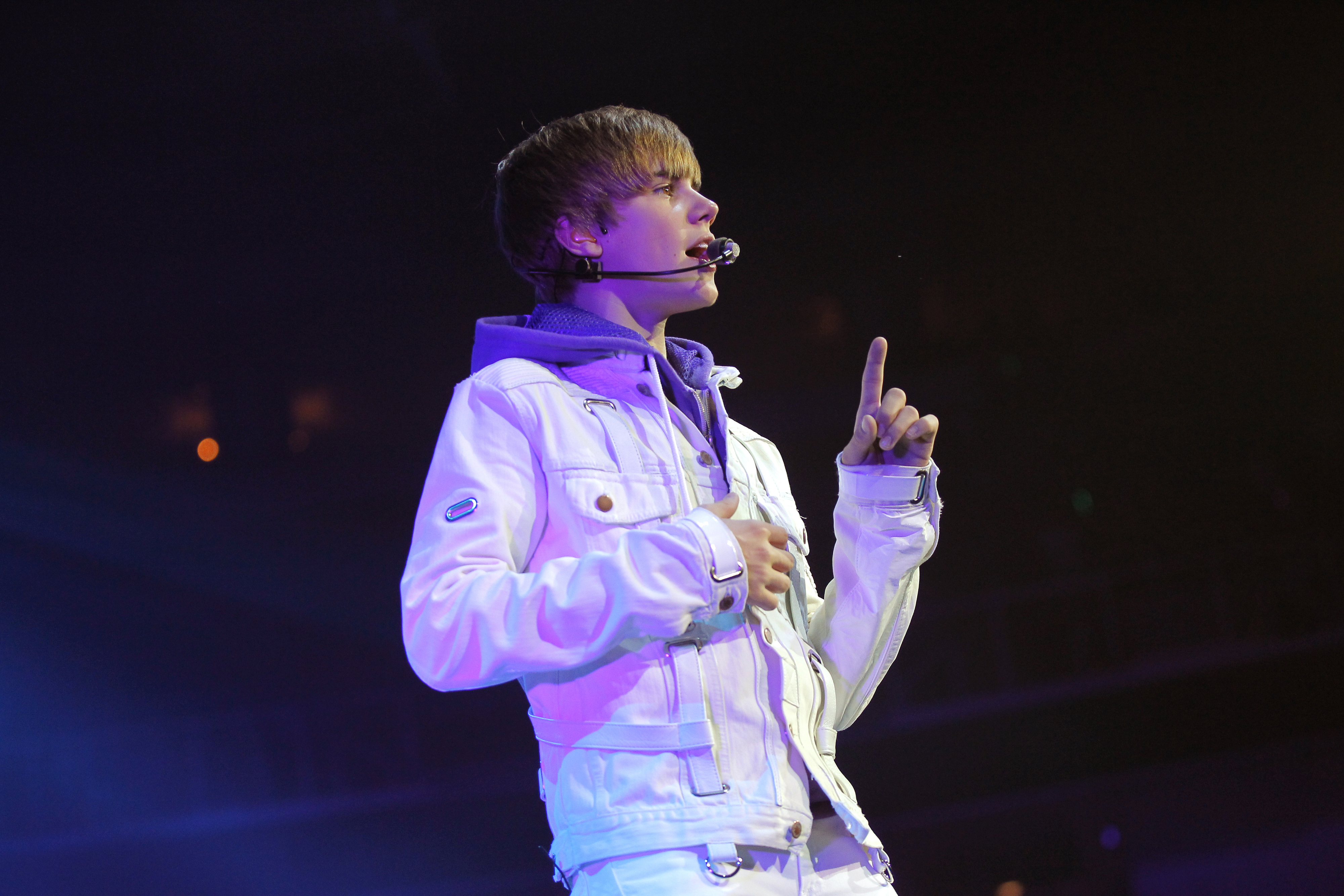 Justin Bieber performing on stage, wearing a casual jacket and headset microphone, gesturing with one hand