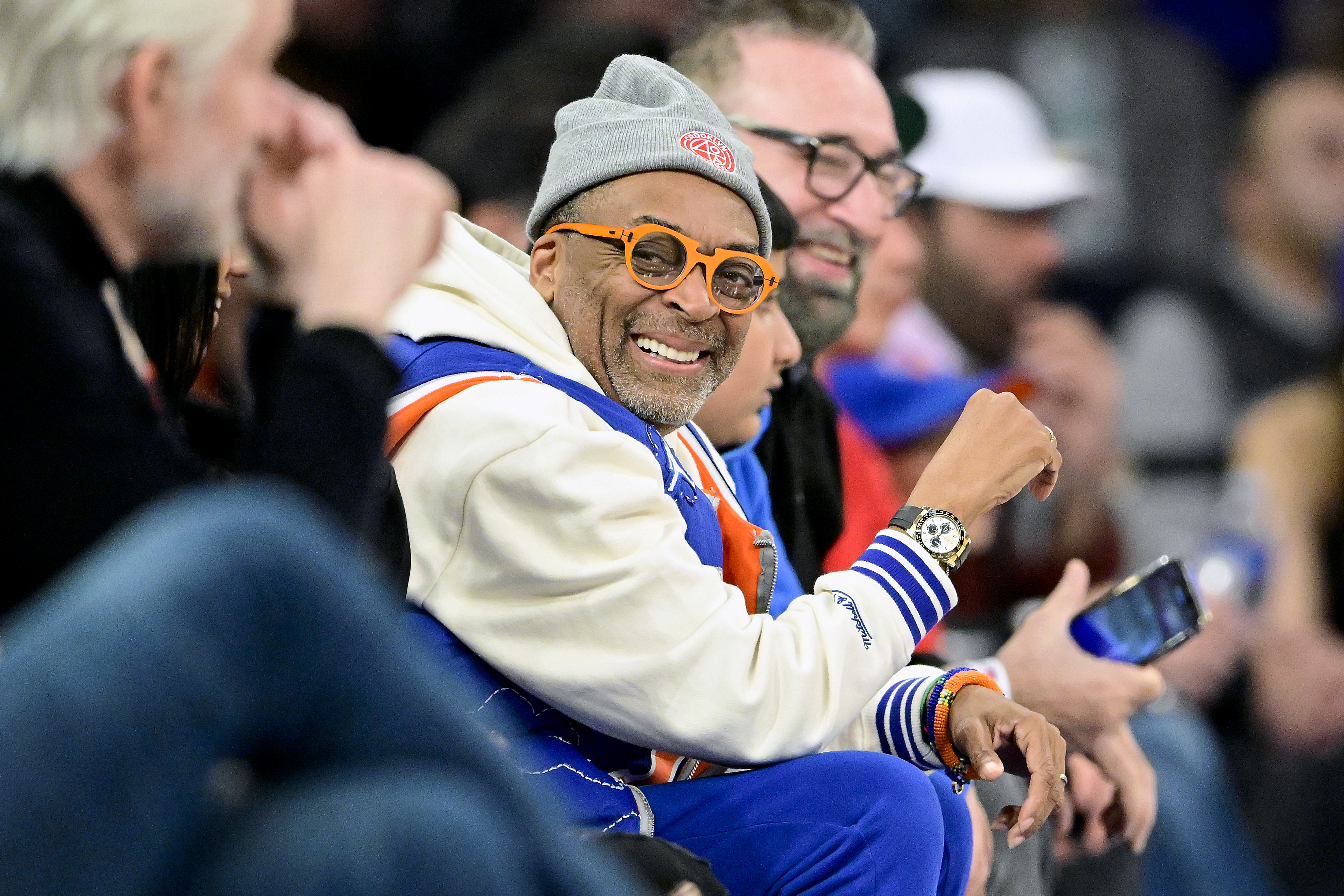 Spike Lee is wearing a white beanie, round glasses, and an orange-and-blue ensemble at a sports event