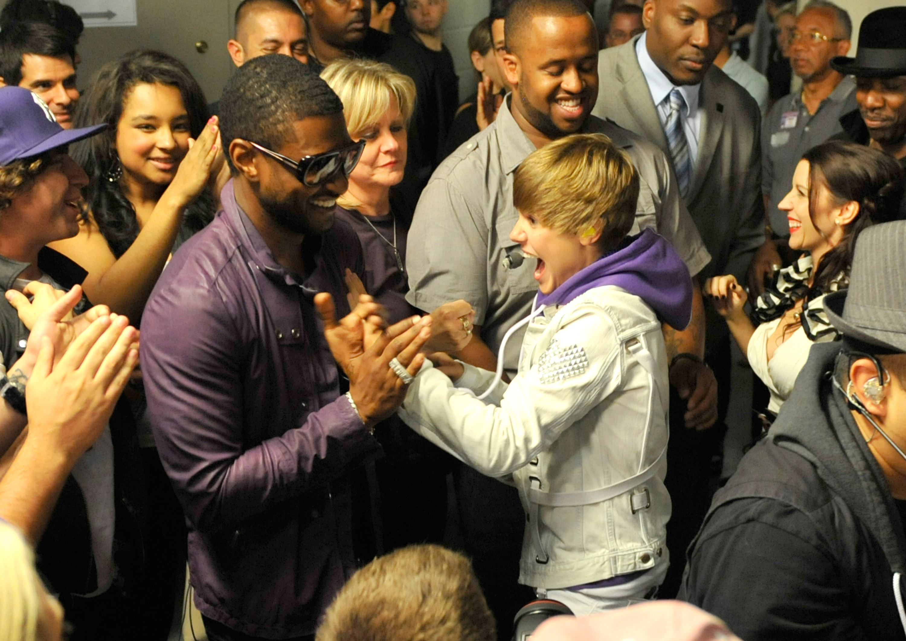 Usher and Justin Bieber shaking hands excitingly in the middle of a crowd backstage