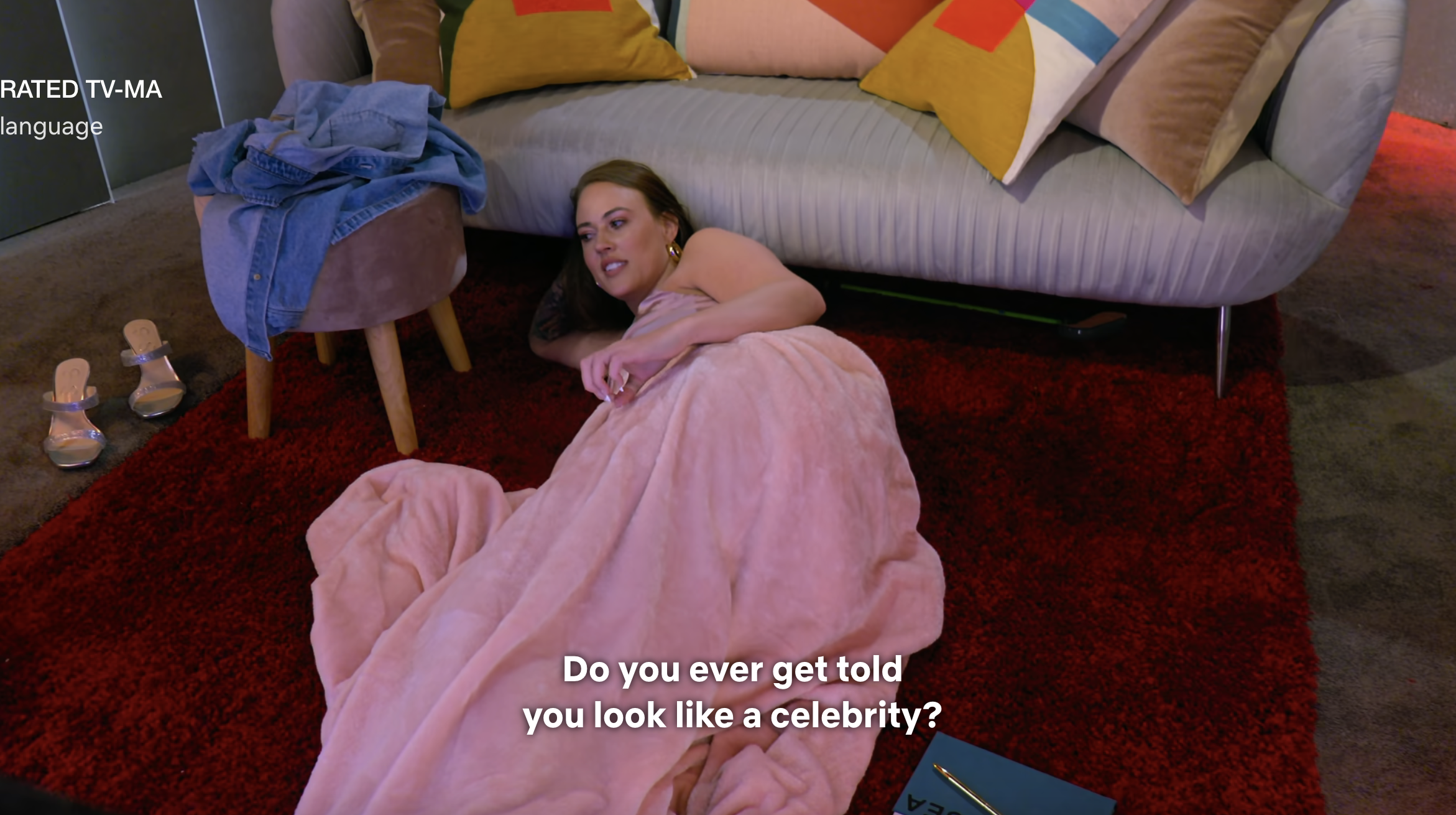 A person lounges on a couch, wearing a flowing pink outfit, with subtitle text &quot;Do you ever get told you look like a celebrity?&quot;