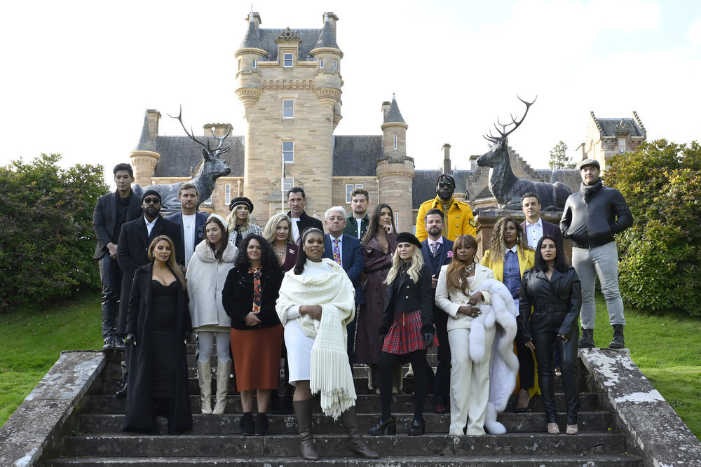 Cast of a TV show standing in front of a castle, dressed in a mix of formal and casual attire