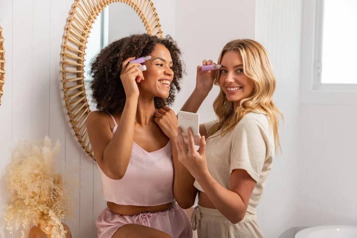 Two women smiling, applying beauty products, in a bathroom setup for a cosmetics ad