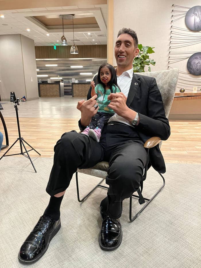 Man in a suit holding a small, lifelike doll, both seated and gesturing &#x27;peace&#x27; signs