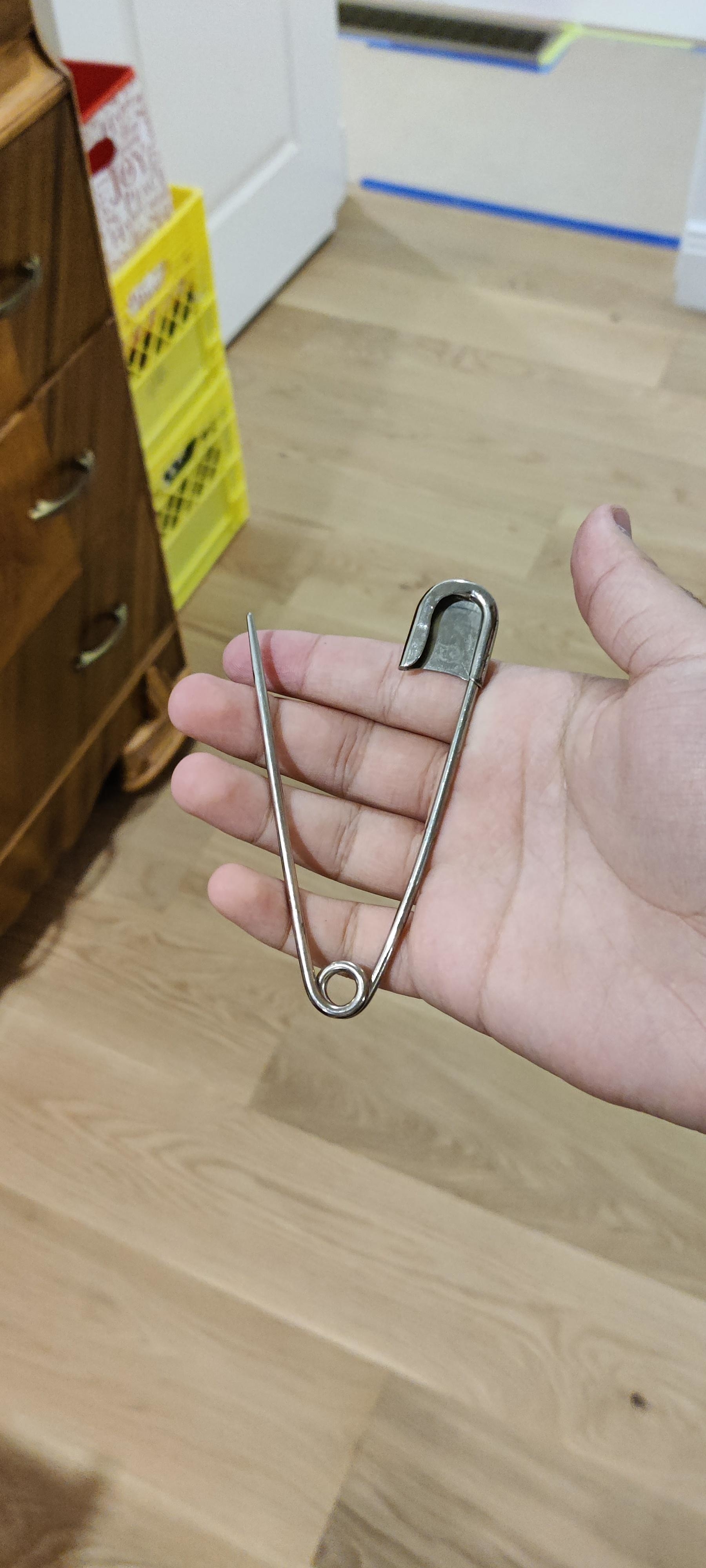 A person&#x27;s hand holding a large metal safety pin against a home interior background