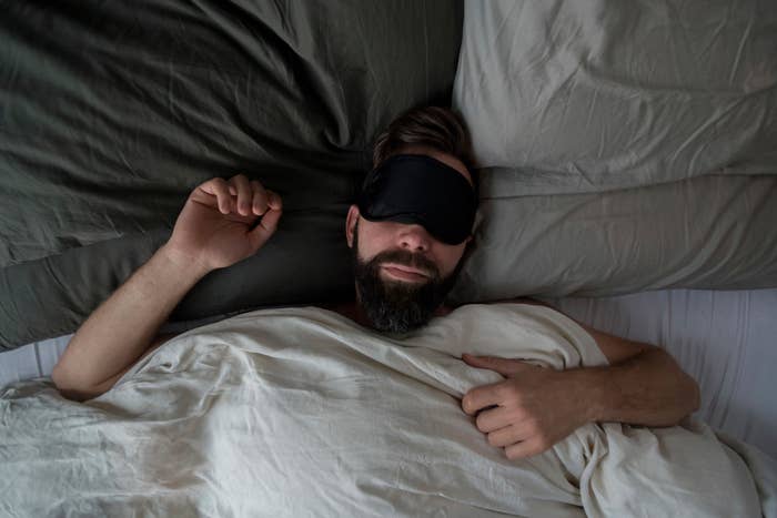 Person wearing eye mask sleeping in bed, with blankets and one arm above head