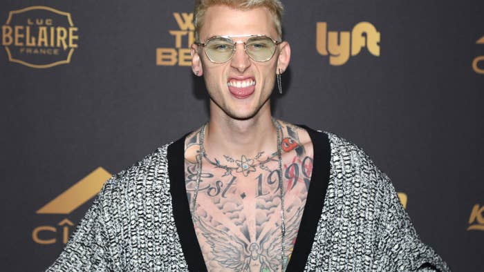 Man in glasses and open patterned jacket revealing chest tattoos, at event with logo backdrop