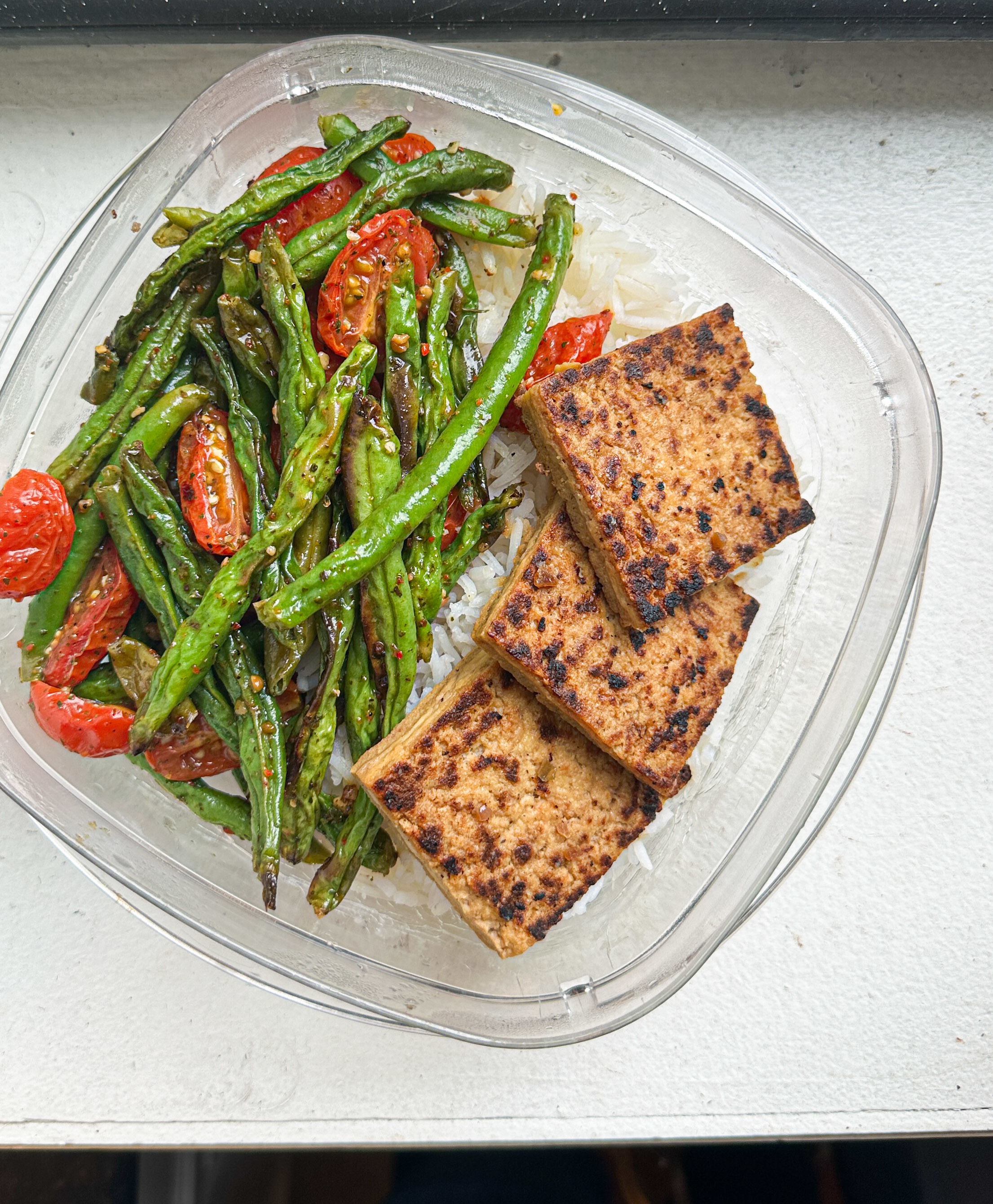 A meal in a clear container with grilled asparagus, cherry tomatoes, and two slices of tofu