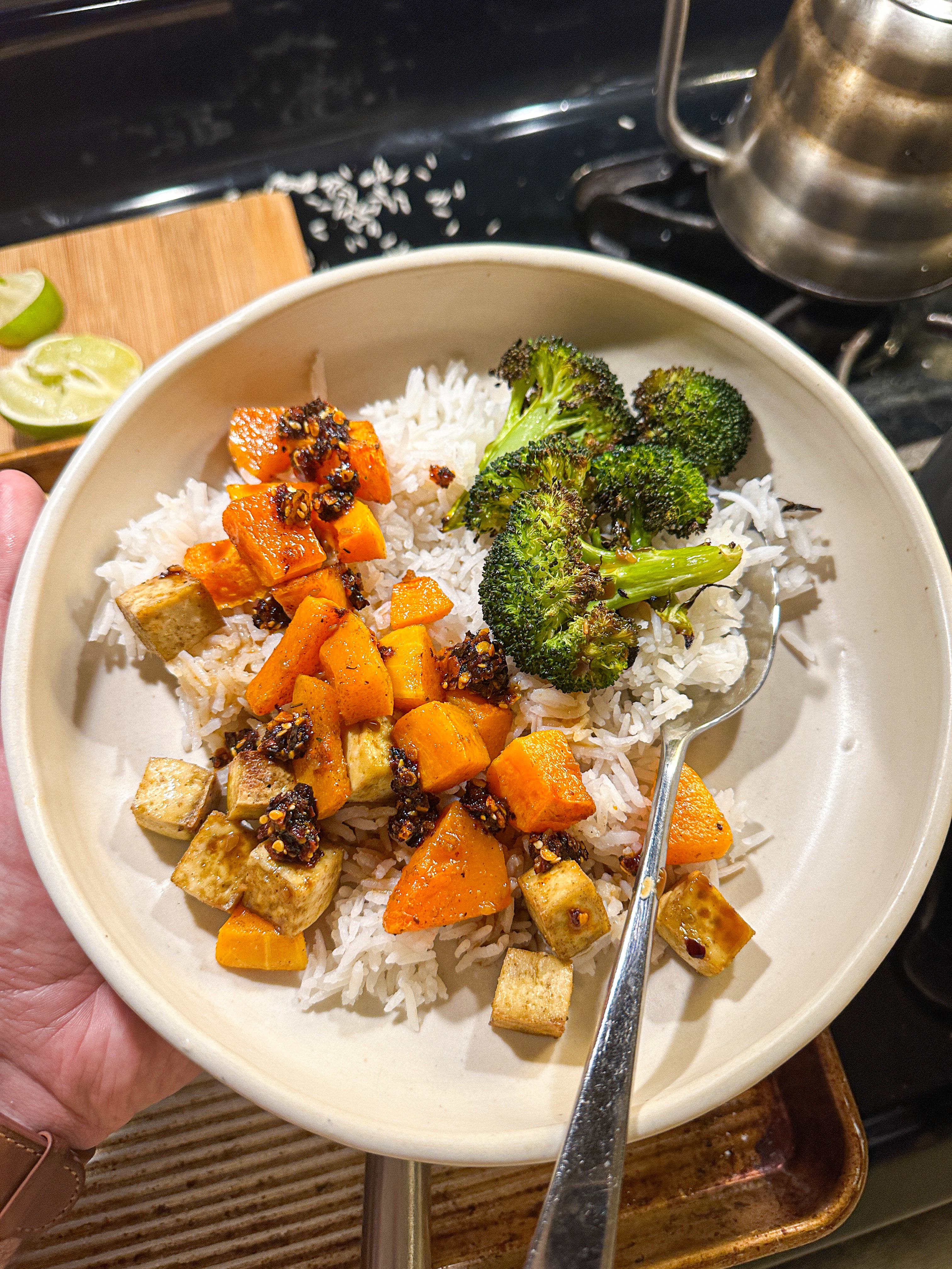 A hand holds a bowl of rice with roasted broccoli, carrots, and tofu cubes, garnished with seeds and lime wedge