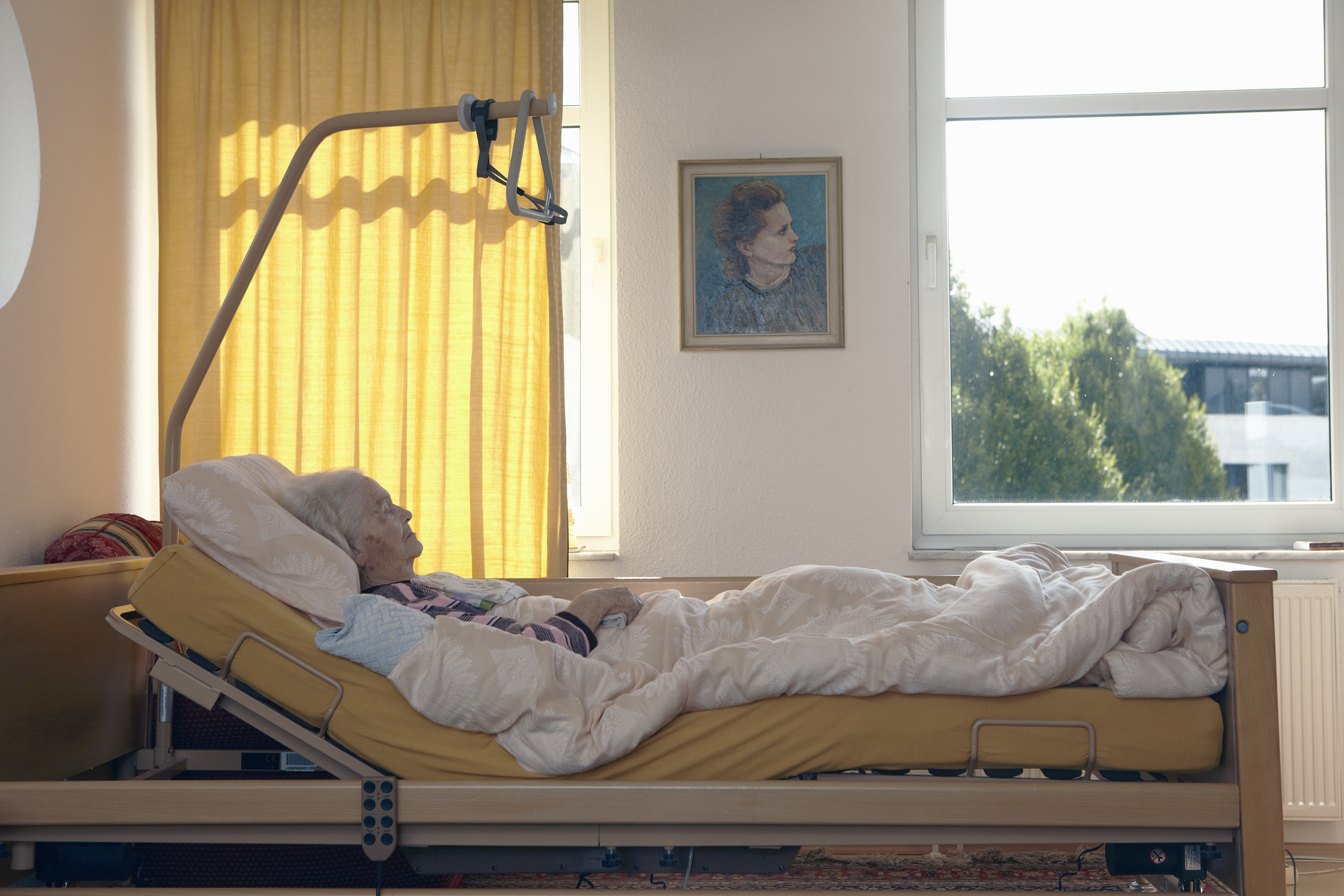 Older woman resting in a hospital bed by a window with a portrait on the wall