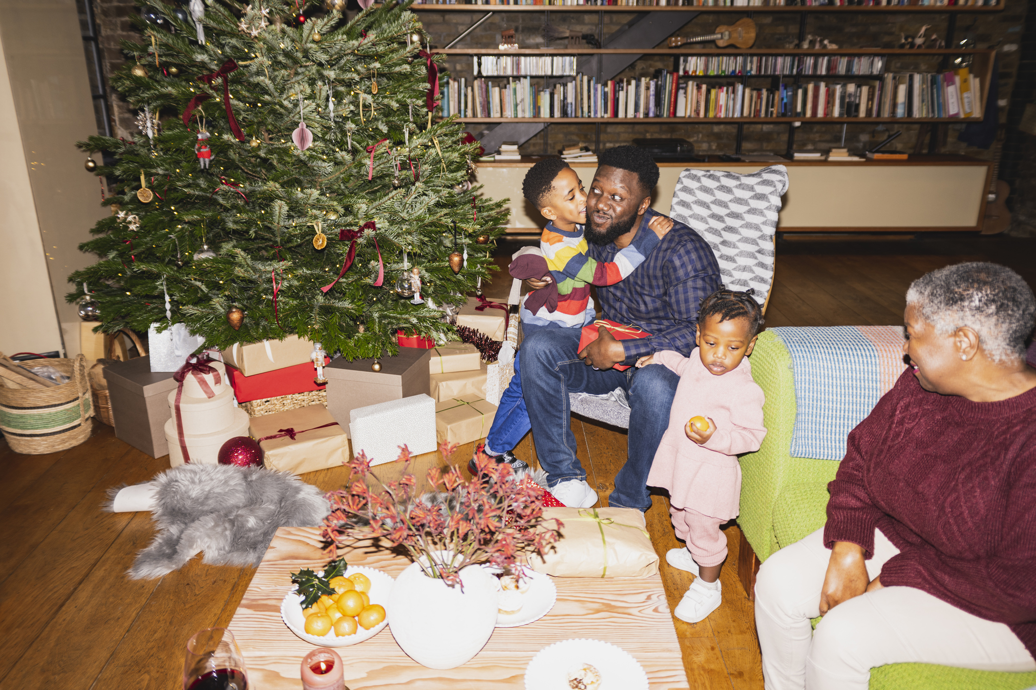 Family with two children next to a Christmas tree, sharing a moment, with presents and a decorated table in the foreground
