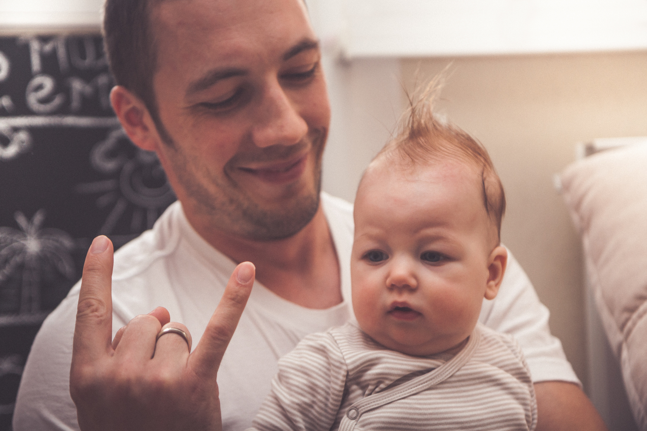 Man holding baby, both looking forward, man makes hand gesture with fingers separated