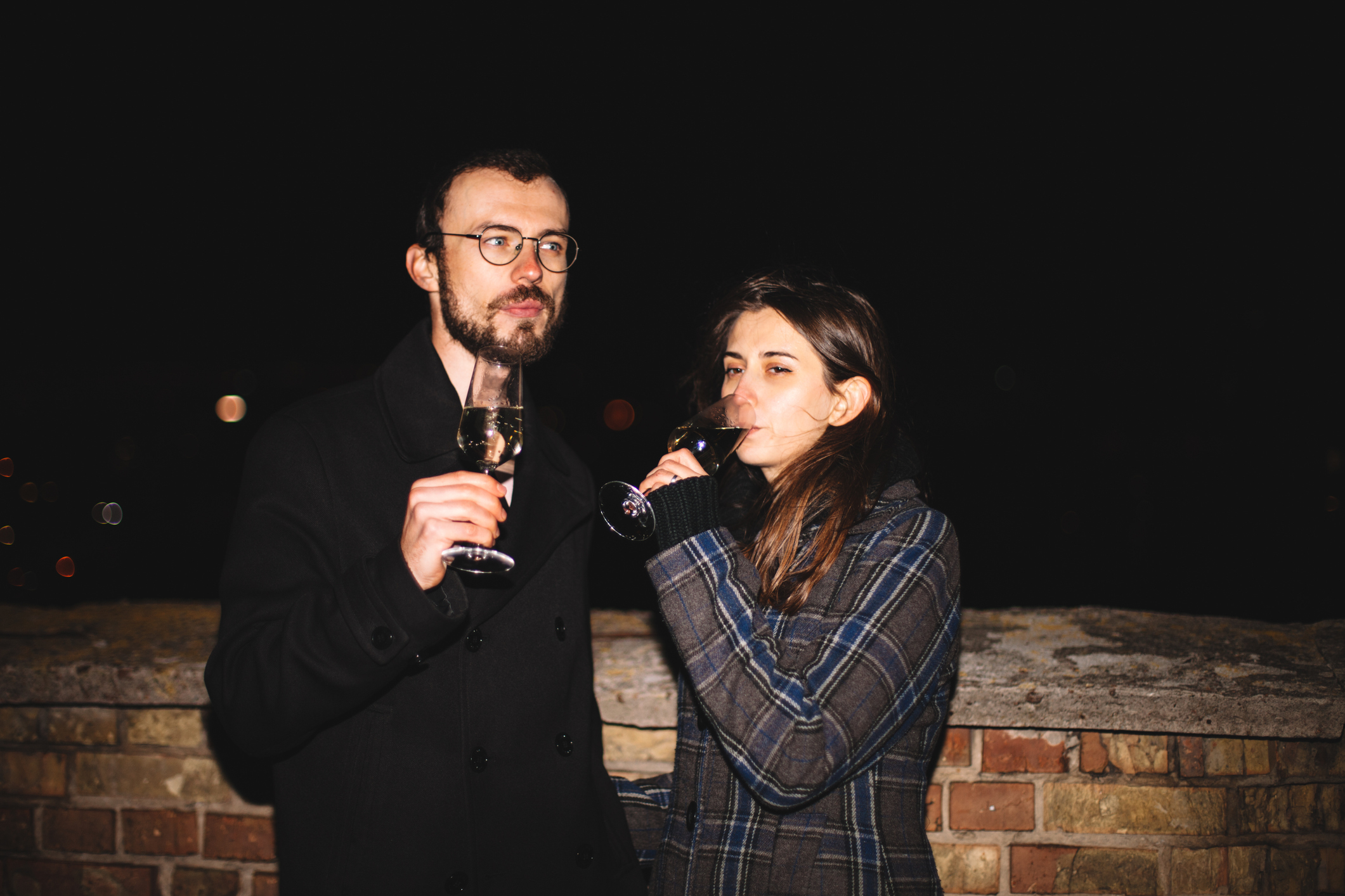 Two people standing close, holding and sipping from wine glasses at night