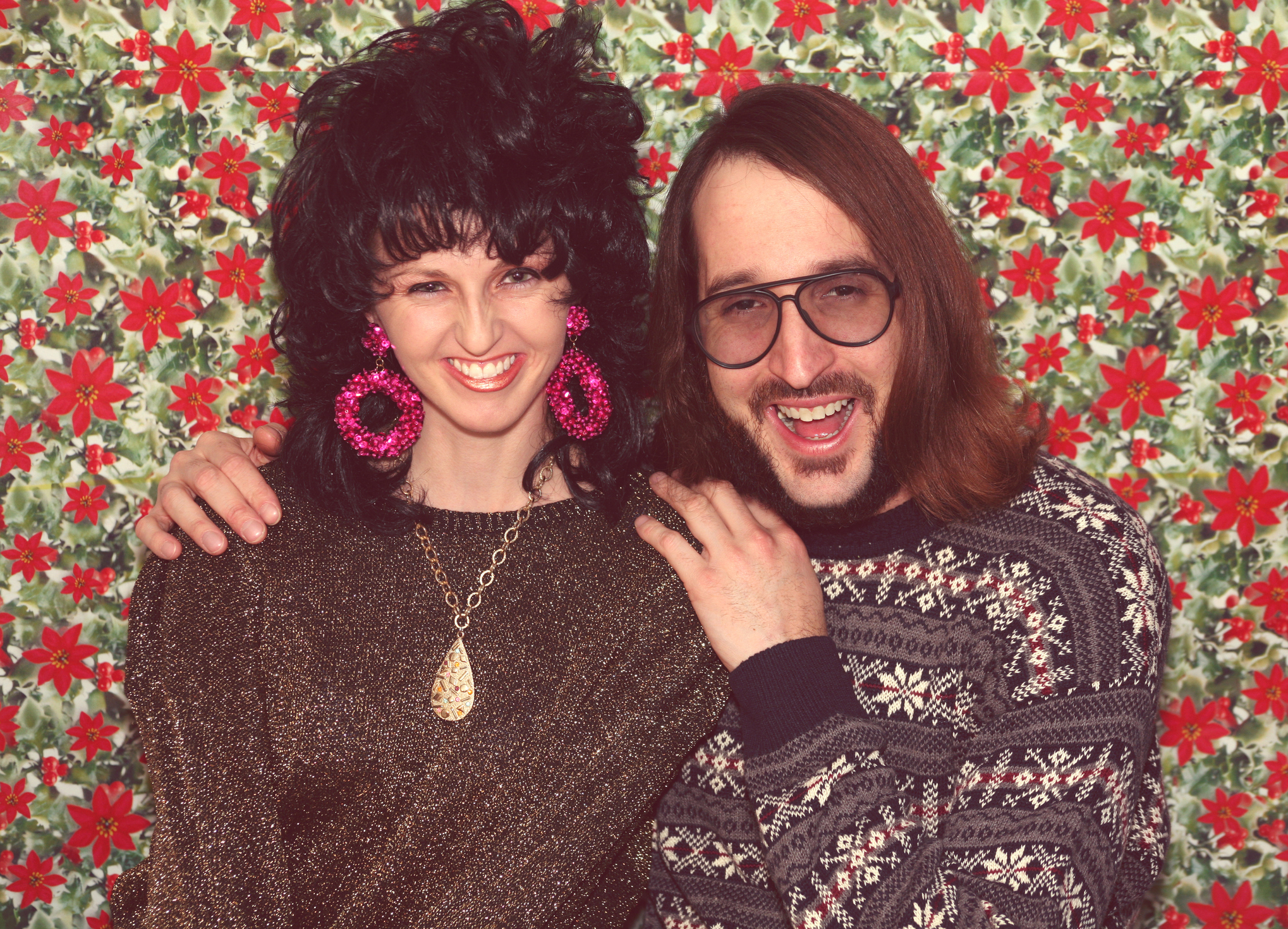 Two people smiling, dressed in vintage style with patterned sweaters, standing before a floral backdrop