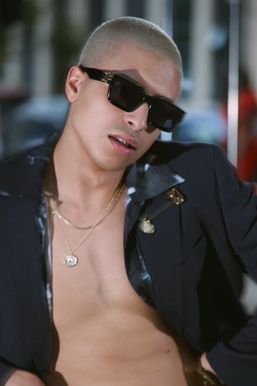 Person with buzz cut wearing sunglasses and an open jacket with no shirt, displaying necklaces