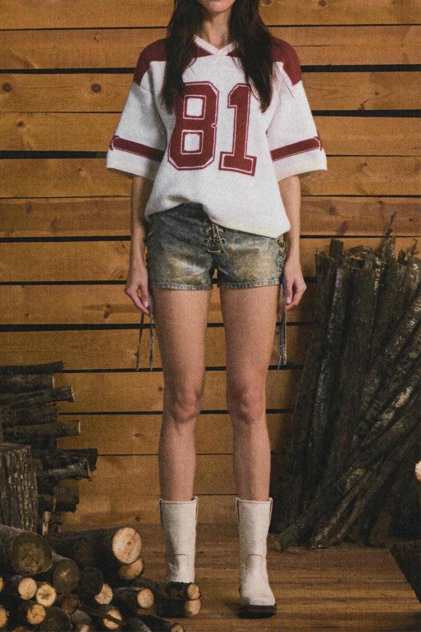 Person in sports jersey and denim shorts with white boots standing in front of a woodpile