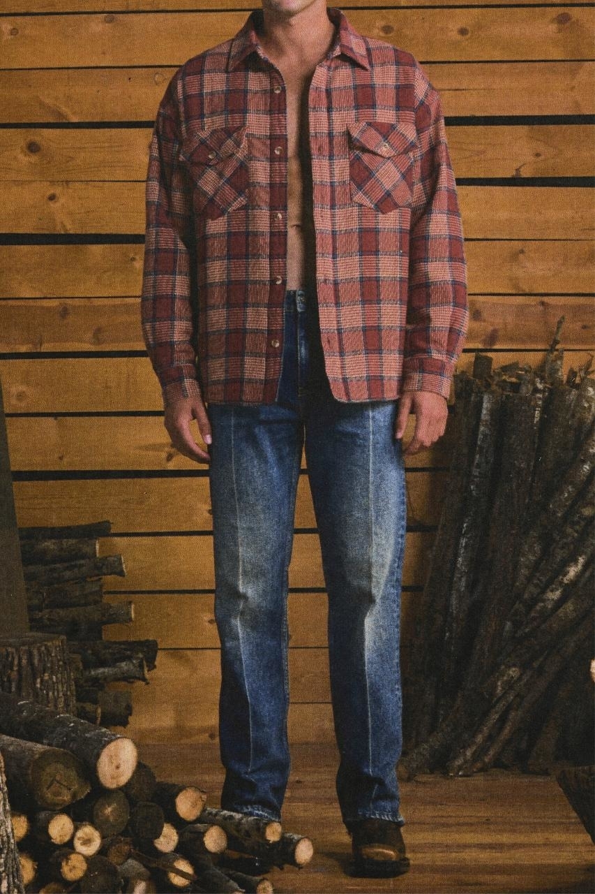 Person in plaid shirt, jeans, and boots standing by firewood stack