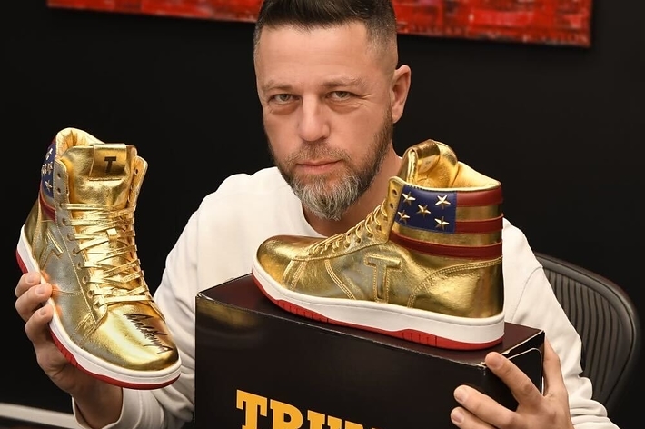 Man holding gold sneakers with 'Trump' on box, sitting at desk