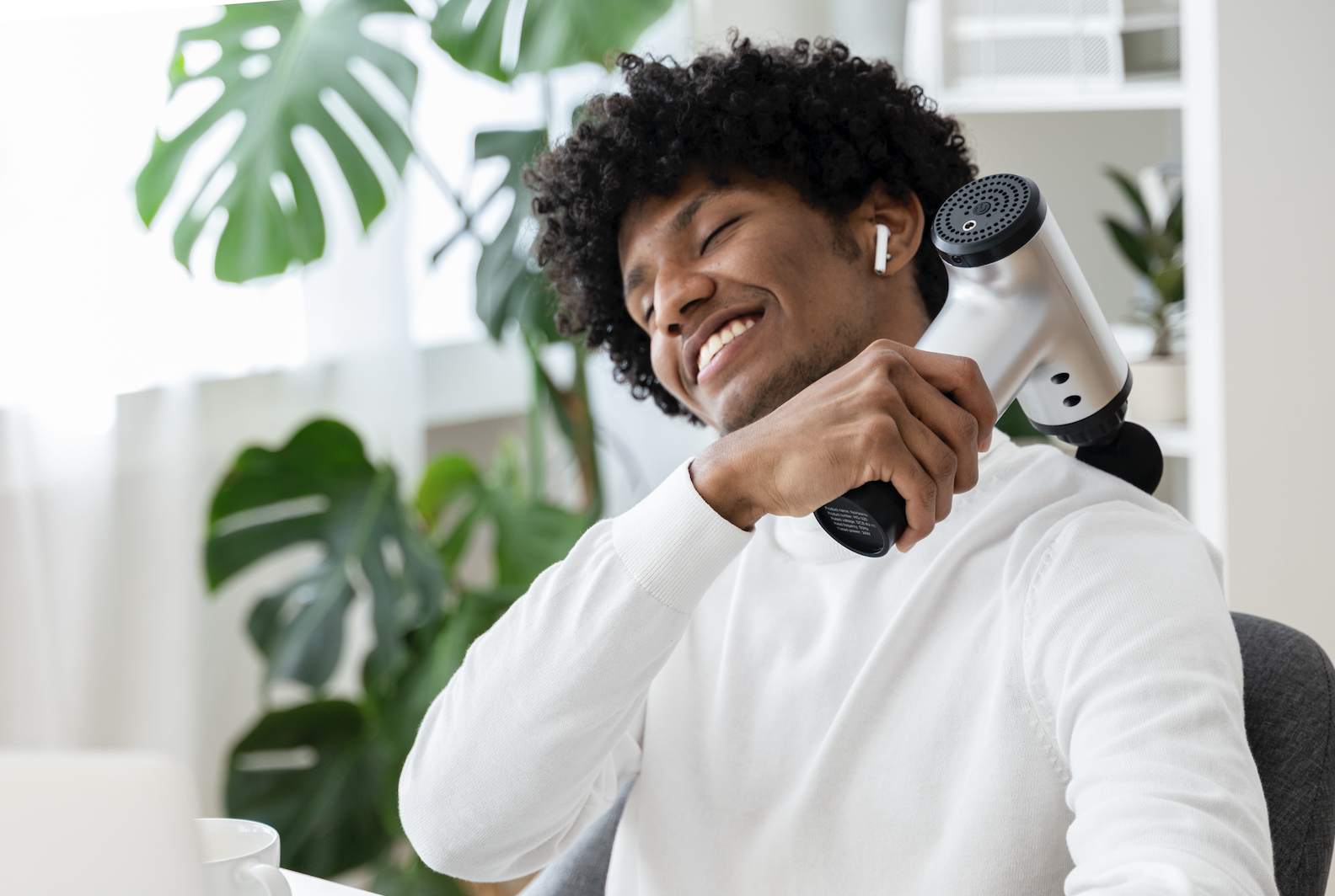 Man using a handheld massager on his neck, eyes closed with a content expression, next to indoor plants