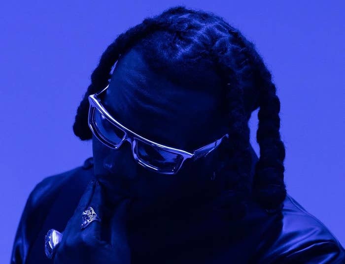 A silhouette of a musician with braided hair and sunglasses, posing thoughtfully against a blue backdrop