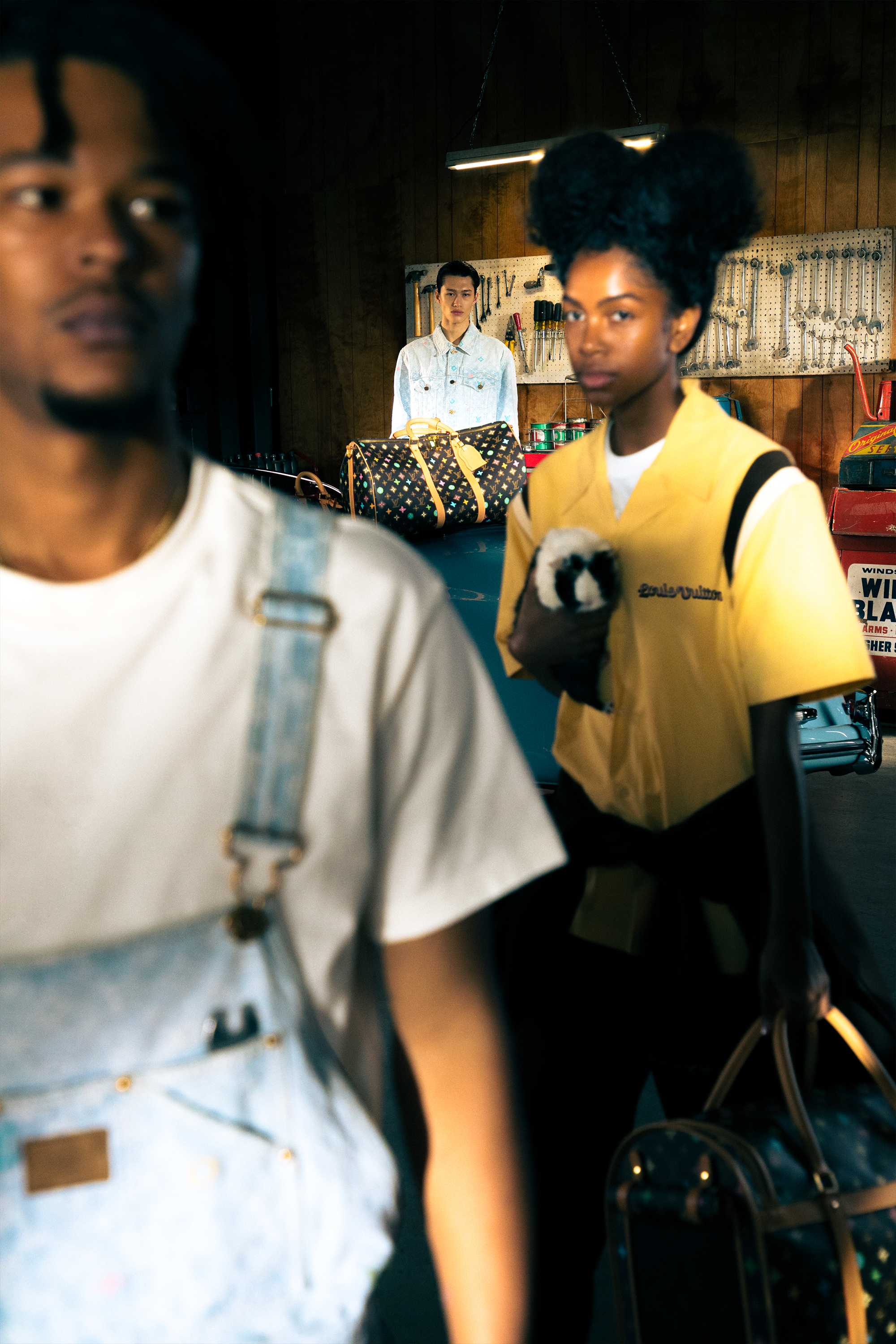 Foreground: Person in overalls with focus on logo. Background: Two people, one holding LV bag