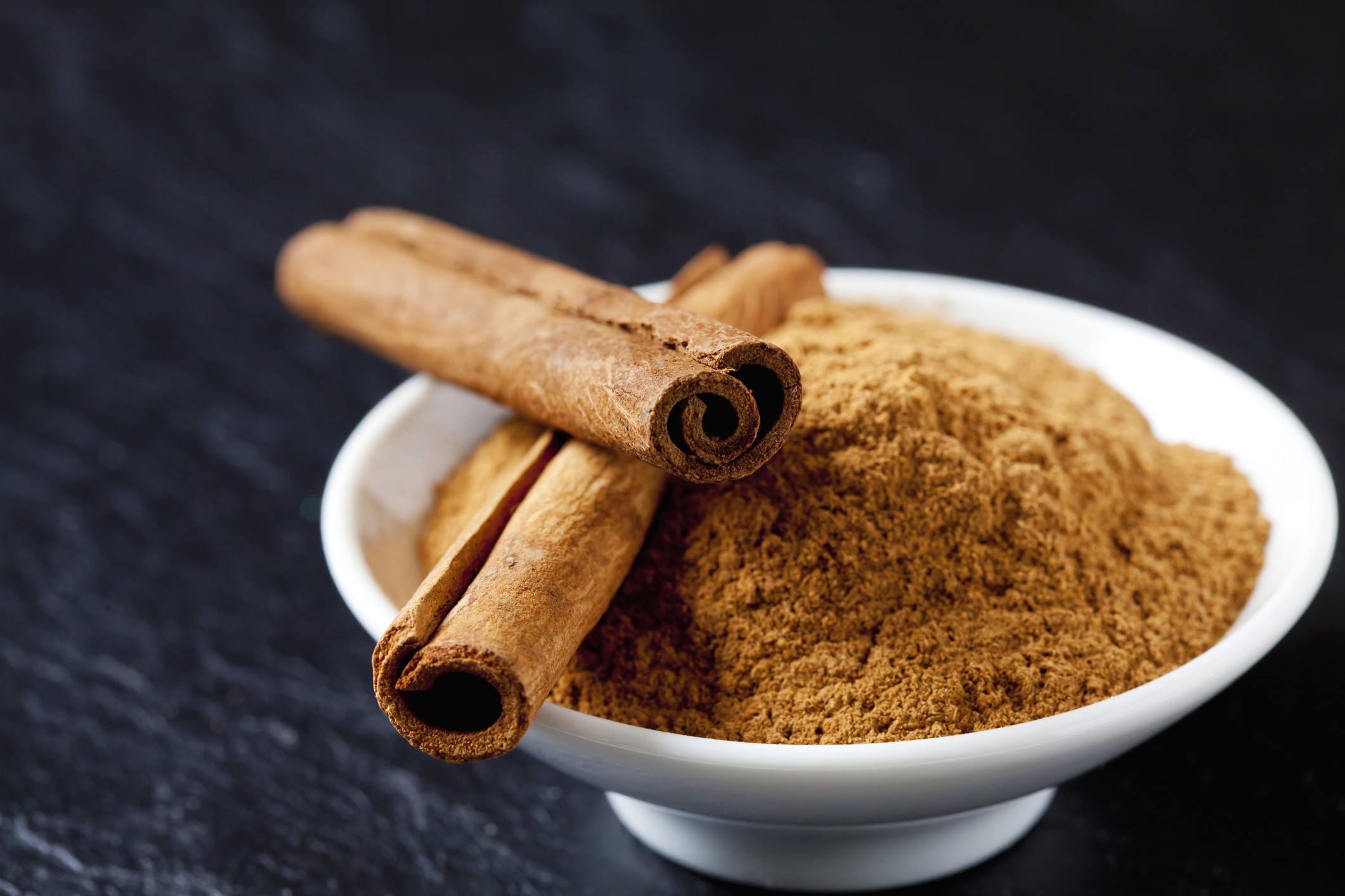 Ground cinnamon and cinnamon sticks in a white bowl on a dark surface