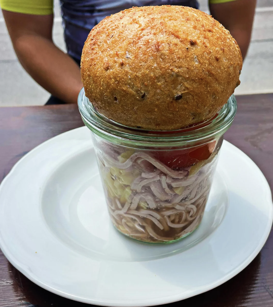 A fried dough ball atop a jar filled with noodles and vegetables, placed on a white plate on a wooden table