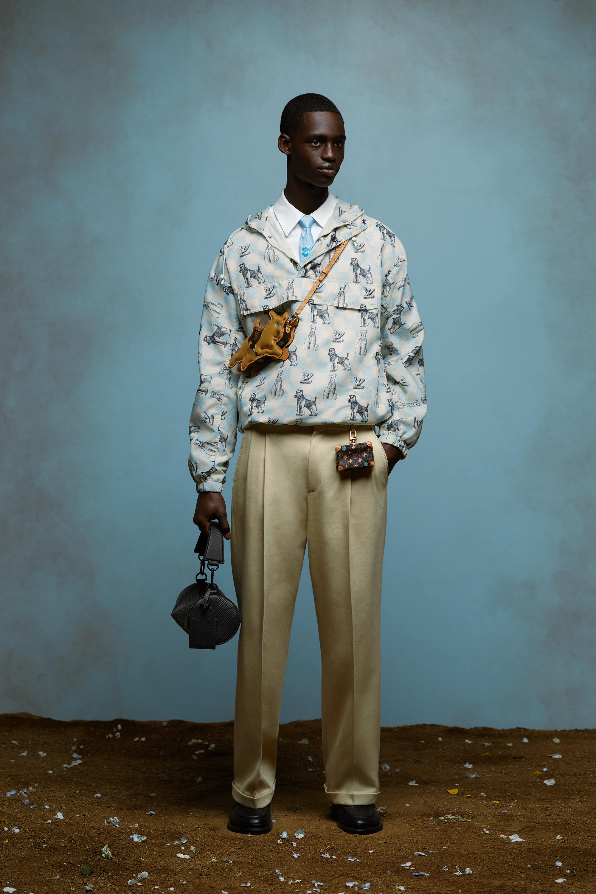 Model in a patterned jacket, layered shirts, beige trousers, and carrying multiple small bags, posing against a blue backdrop