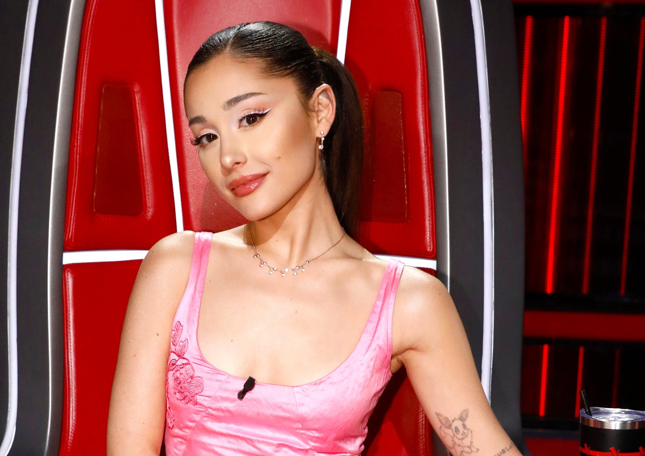 Ariana Grande seated in a coach chair wearing a pink satin dress with a sleek hairstyle