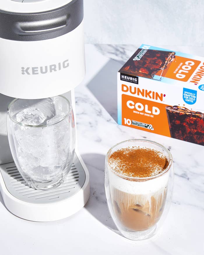 A Keurig machine brewing a cold Dunkin&#x27; coffee into a glass with ice, with Dunkin&#x27; K-Cup box visible