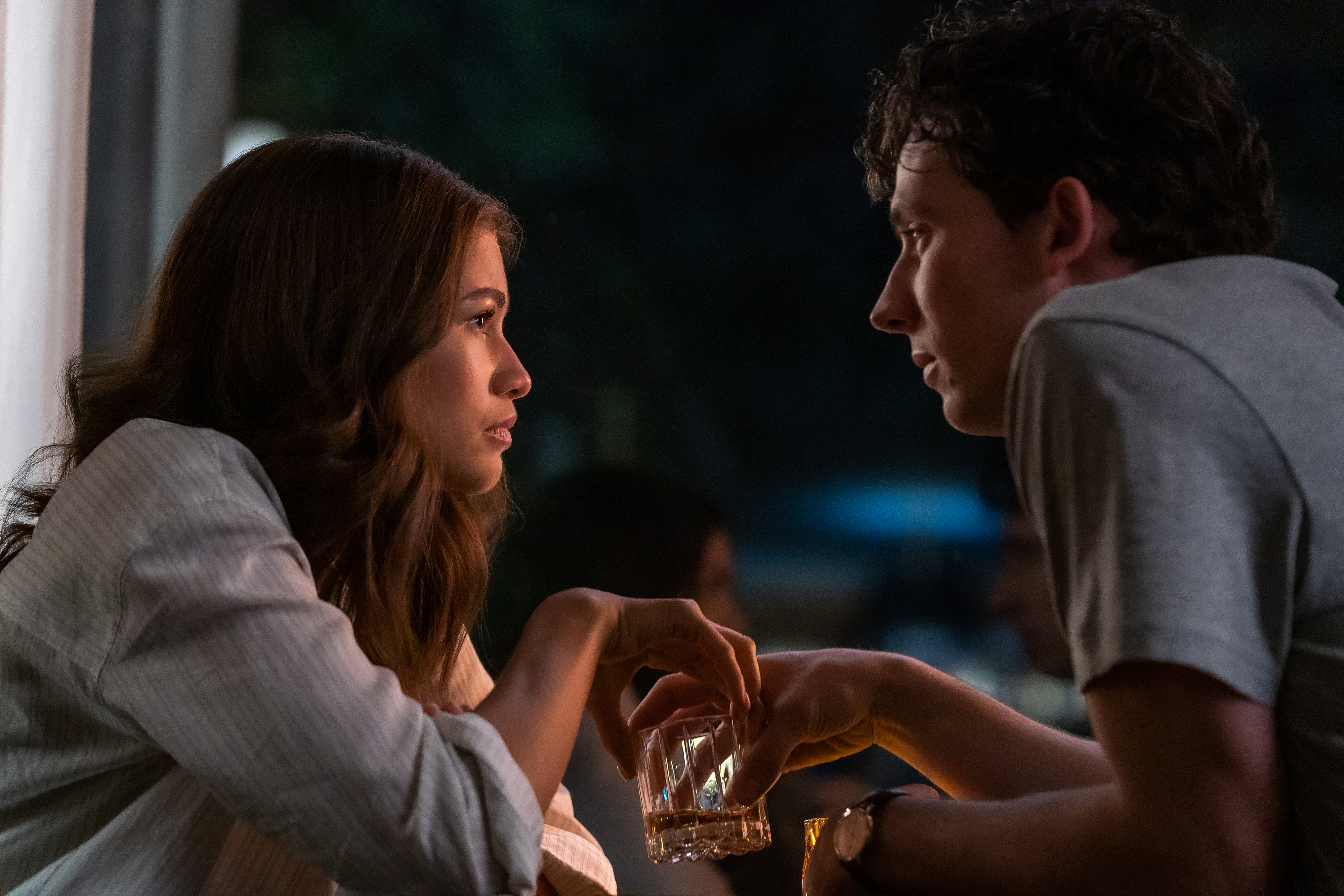 Two characters from a scene are engaged in a close, intense conversation over a drink