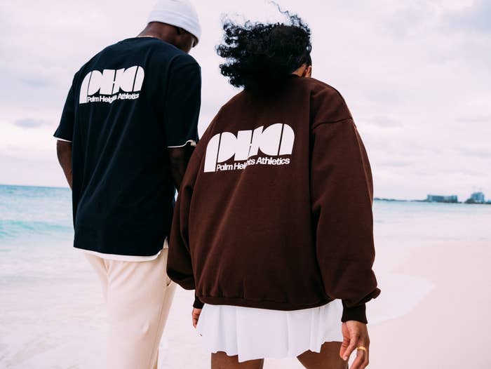Two people on a beach wearing sweatshirts with &quot;Palm Angels Athletica&quot; branding, facing away from the camera