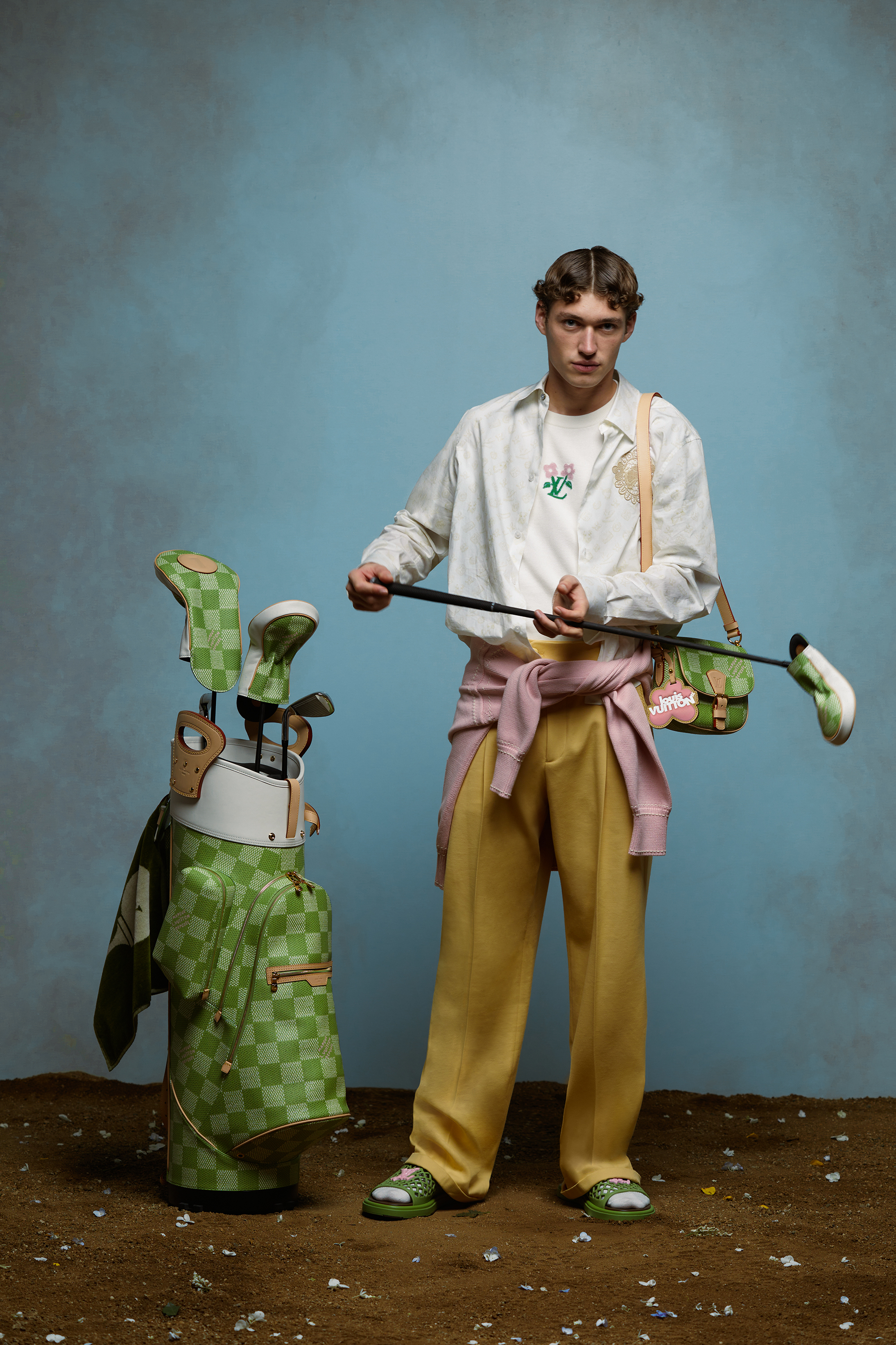 Person in eclectic outfit with golf bag, floral embroidery on shirt, pastel sash, and sandals