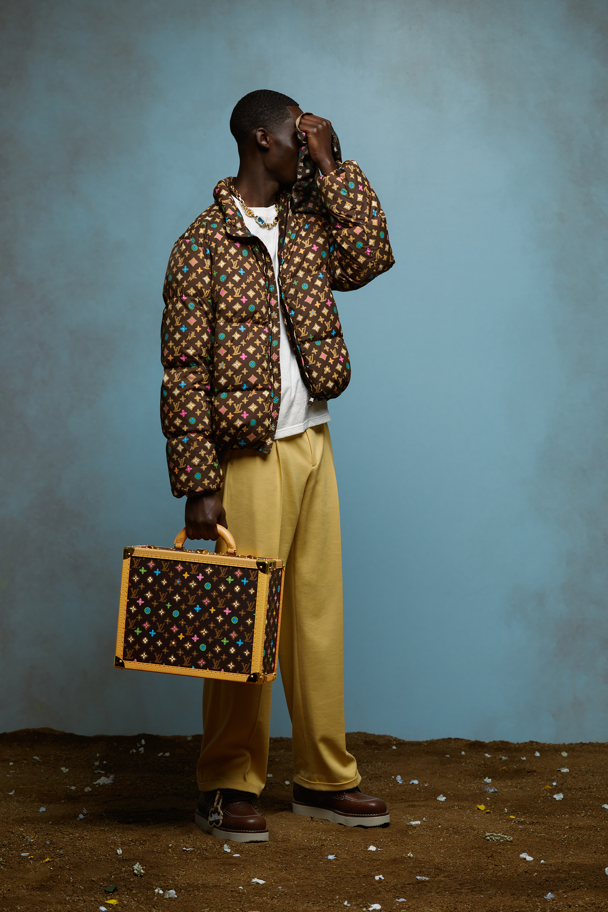 Man in patterned jacket and yellow trousers holding a patterned suitcase, posing in profile