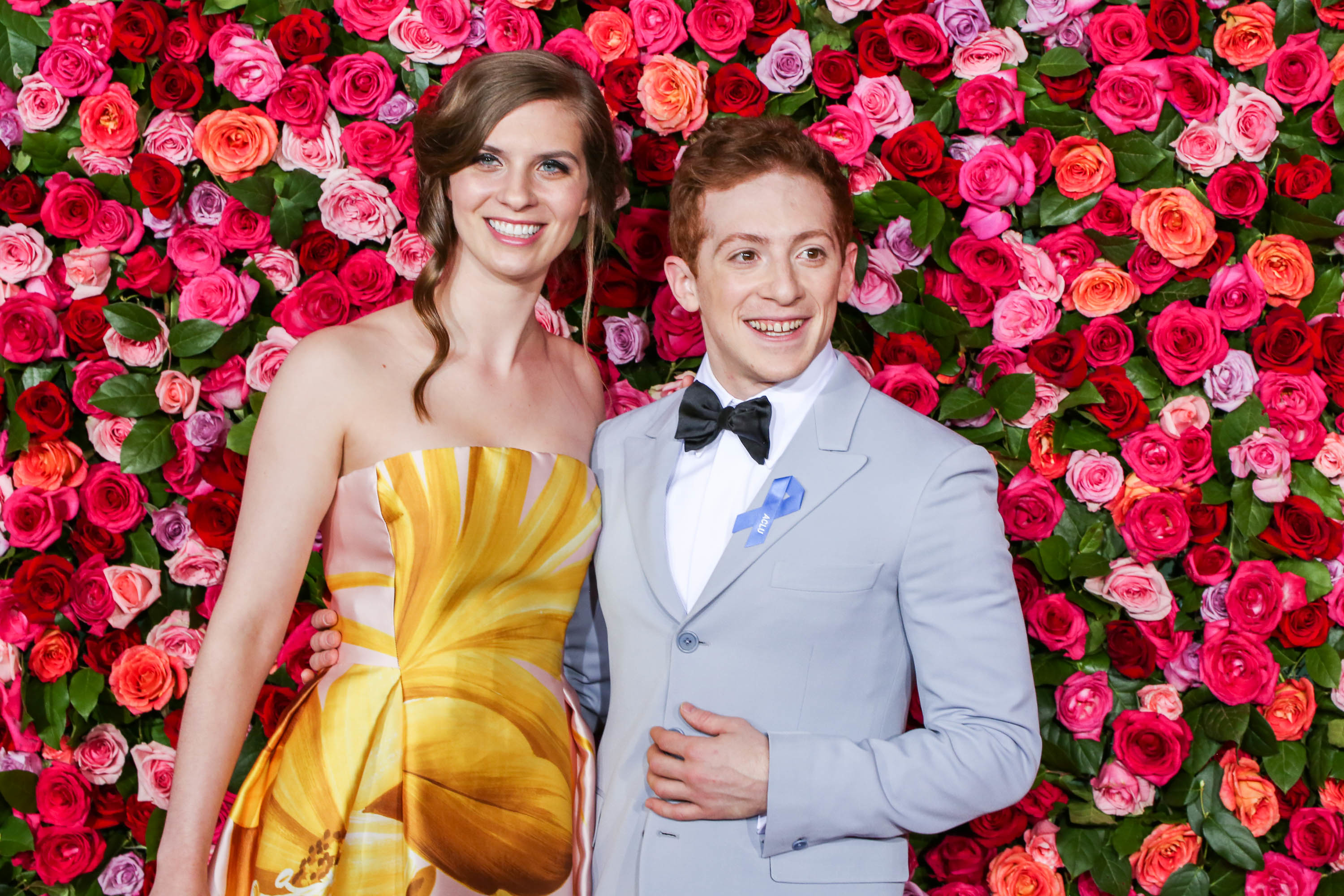 Two people posing in front of a floral backdrop, one in a patterned dress and the other in a light-colored suit