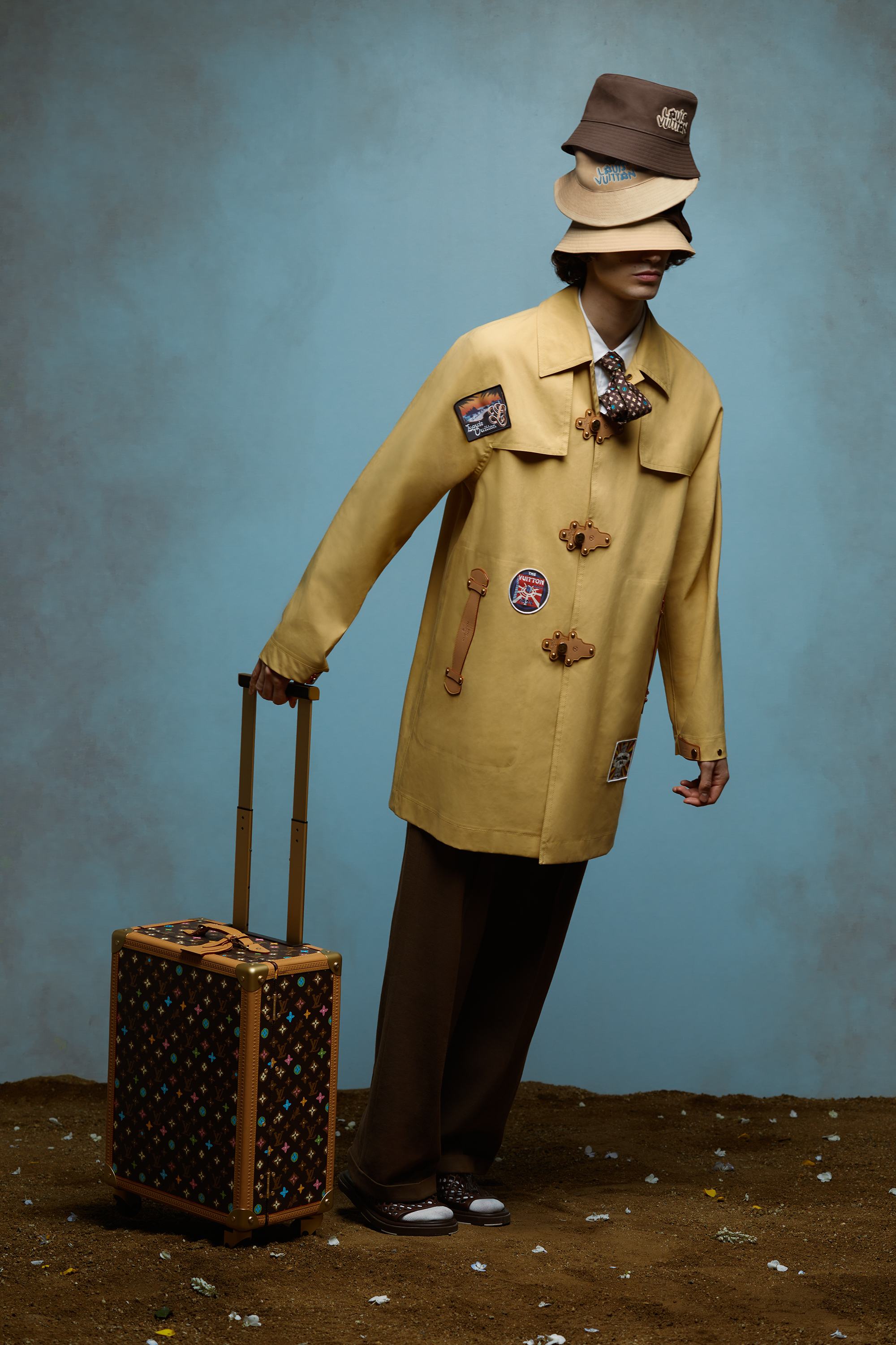 Person in a stylish trench coat with patches, hat, holding a patterned suitcase