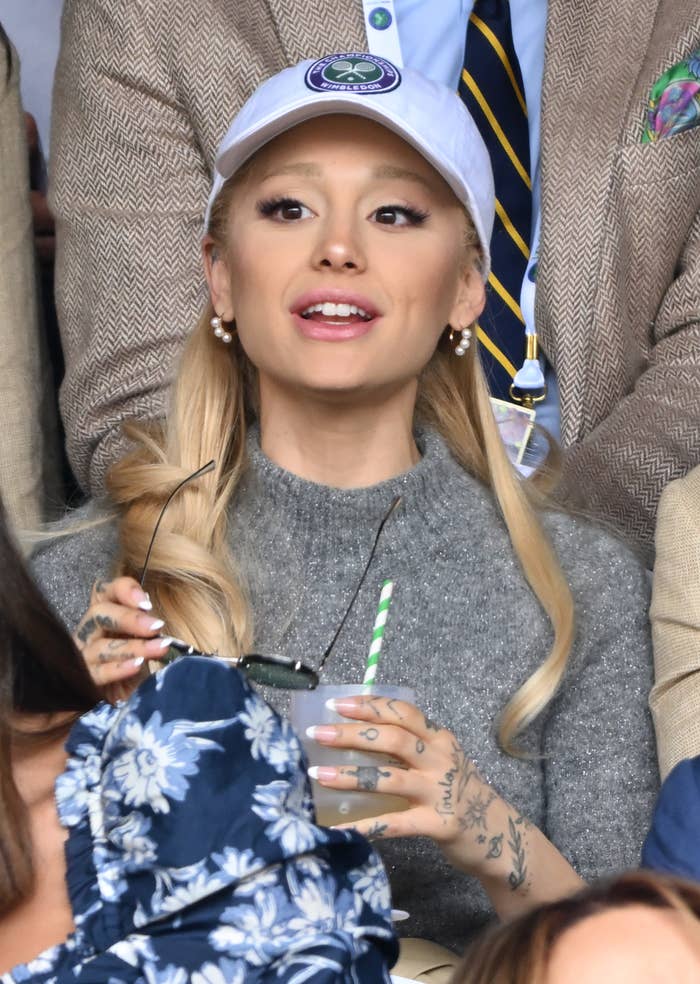 Woman wearing a cap and a grey top at a sports event, holding a drink, with tattoos on her hands
